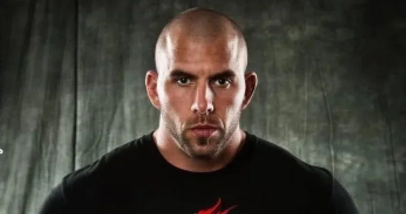 A man with a shaved head and a serious expression, wearing a black shirt, standing in front of a dark, textured background.
