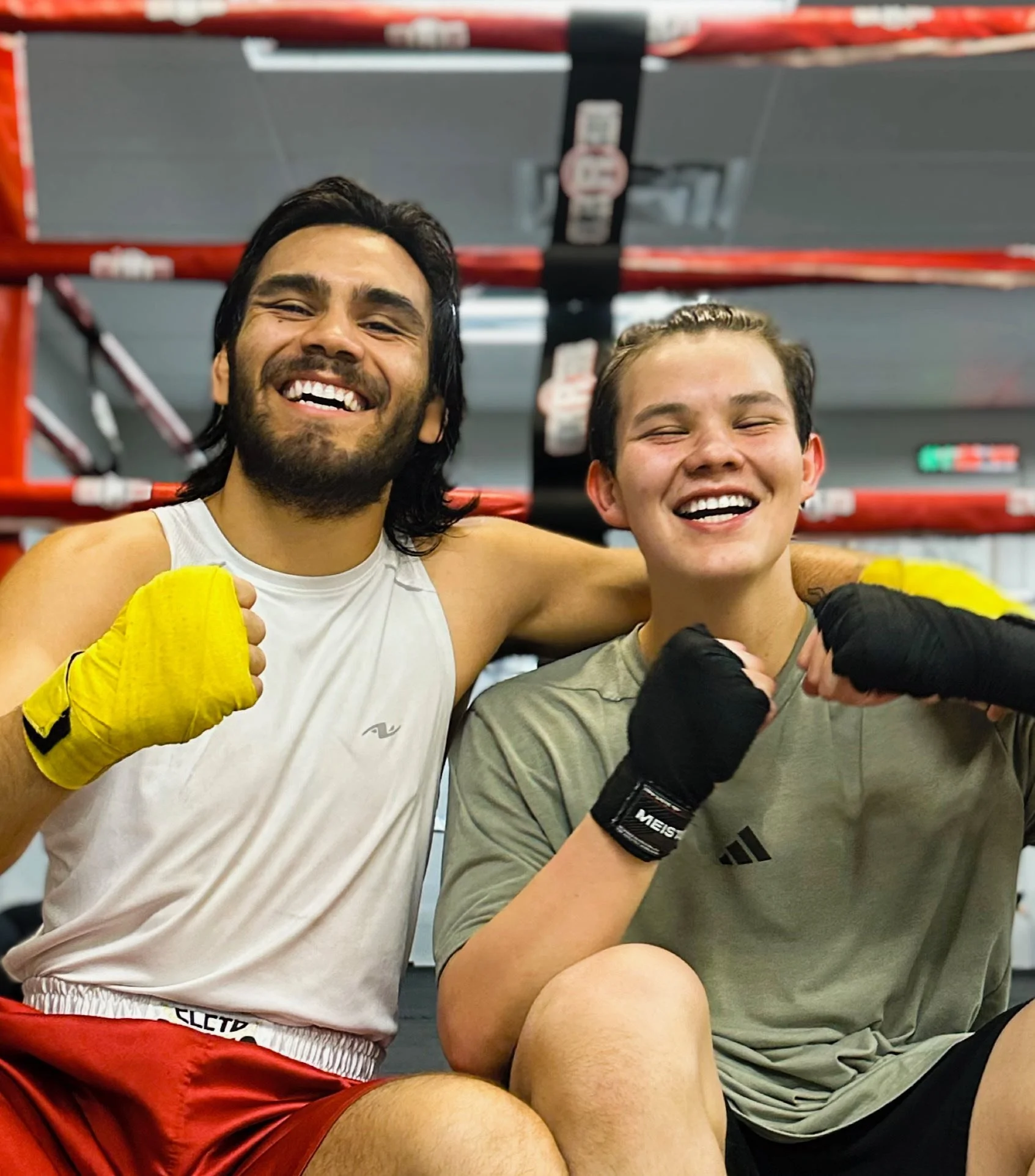 Two young male boxers, smiling and making fists, sitting together after a boxing match in a gym with boxing rings in the background.