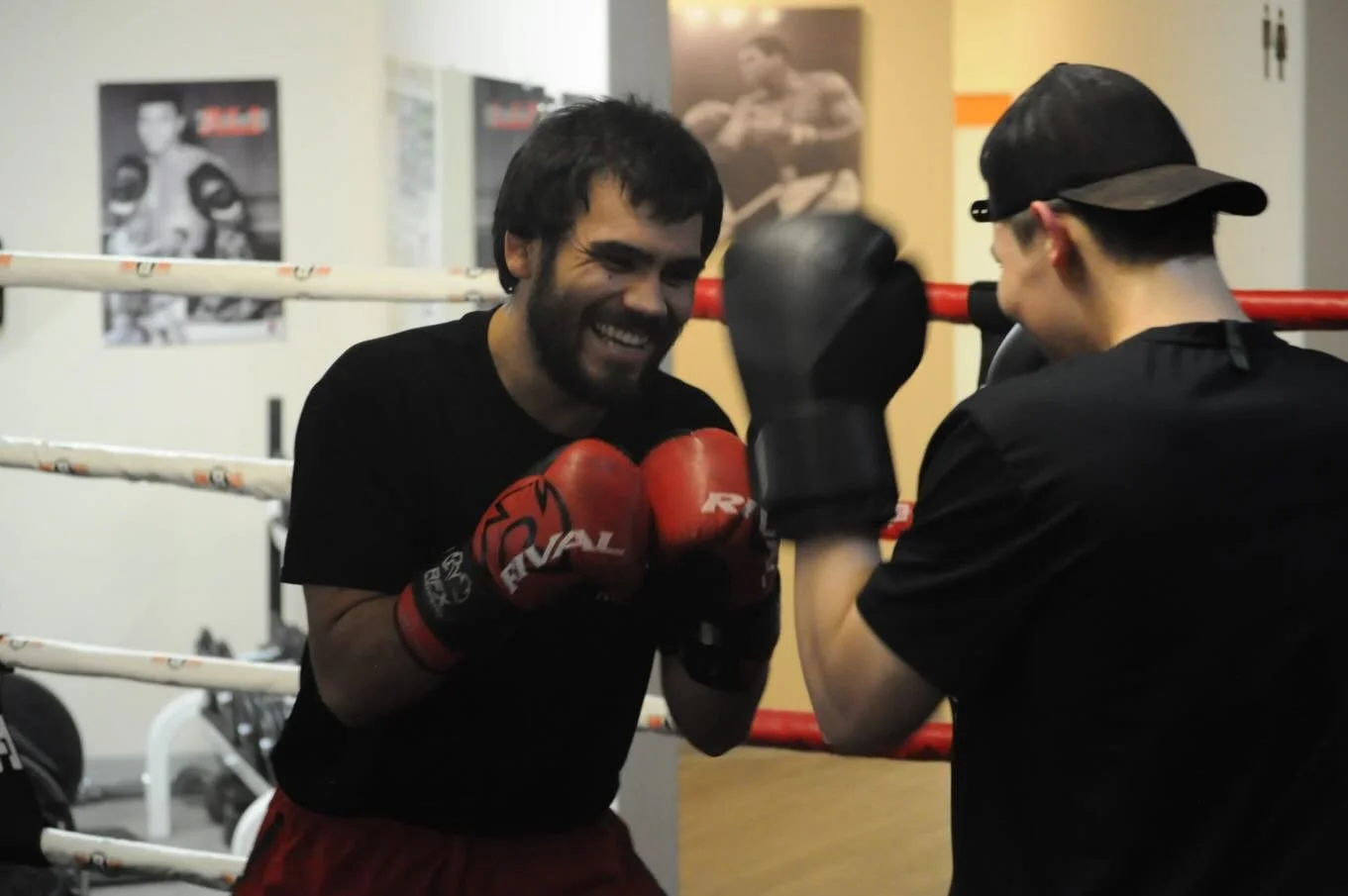 Two men boxing in a gym, smiling and wearing boxing gloves, with one sparring partner and a boxing ring in the background.