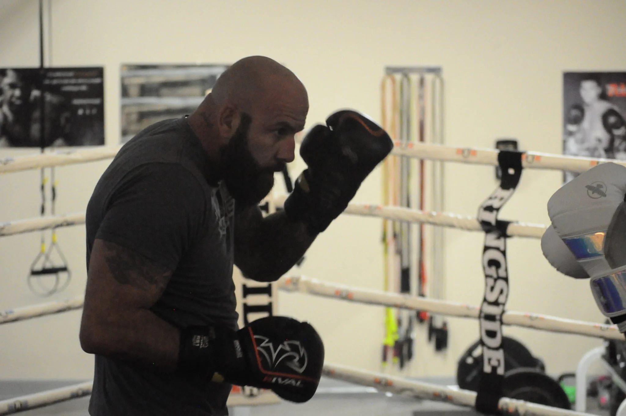 A bald man with a beard in a boxing gym, wearing black gloves, is practicing boxing stances inside a boxing ring.