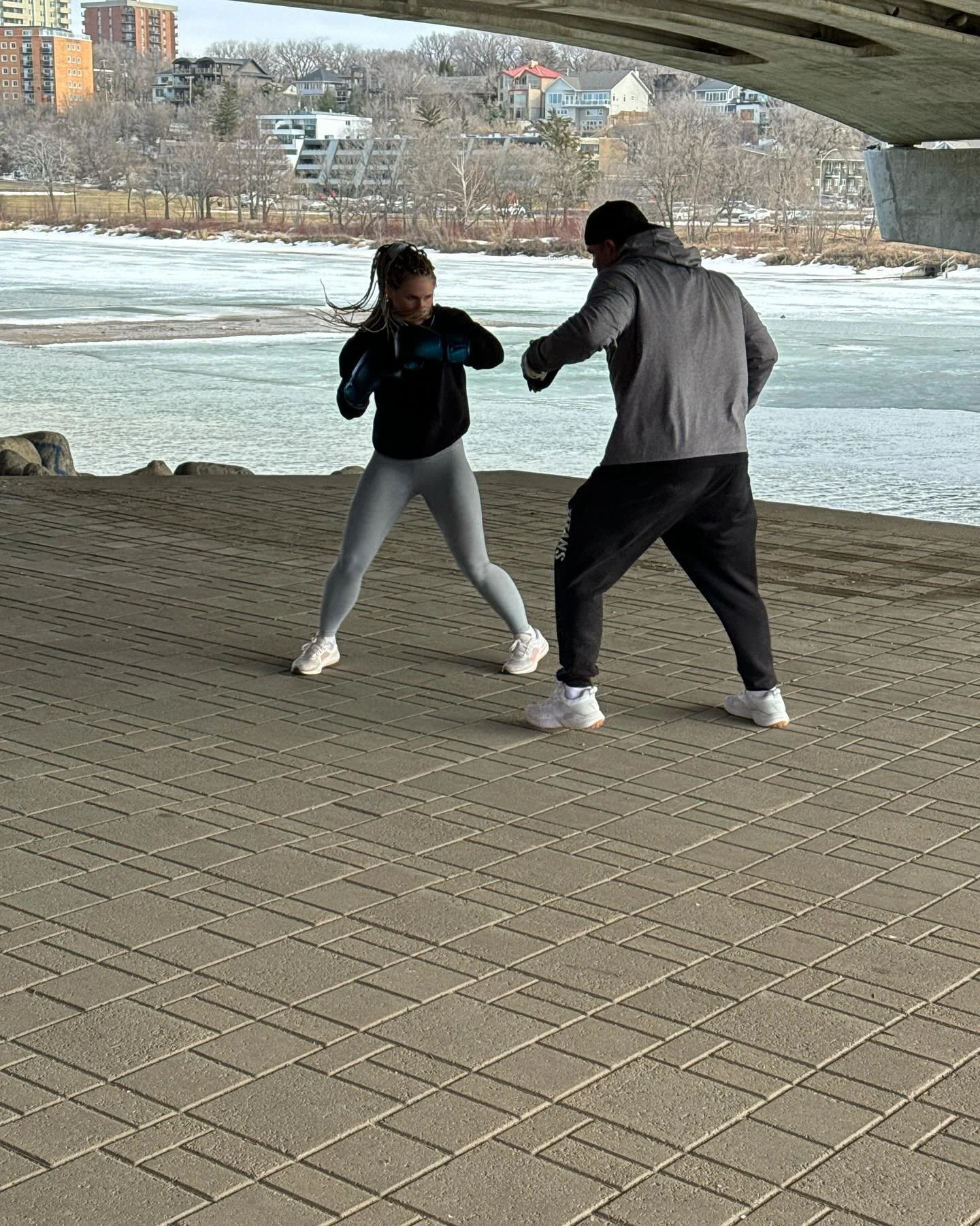 A man and woman practicing boxing outdoors under a bridge near a frozen river with houses and trees in the background.