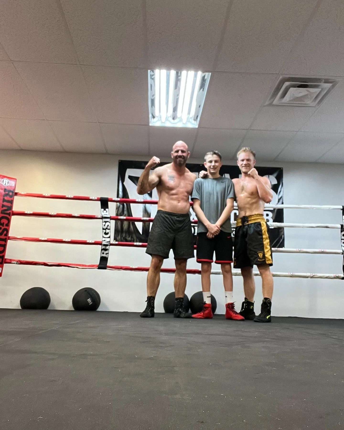 Three people standing inside a boxing gym, two shirtless men flexing their muscles and a young boy with his arms crossed, all smiling. They are in front of a boxing ring with black and red ropes. The gym has a black banner with white text behind them