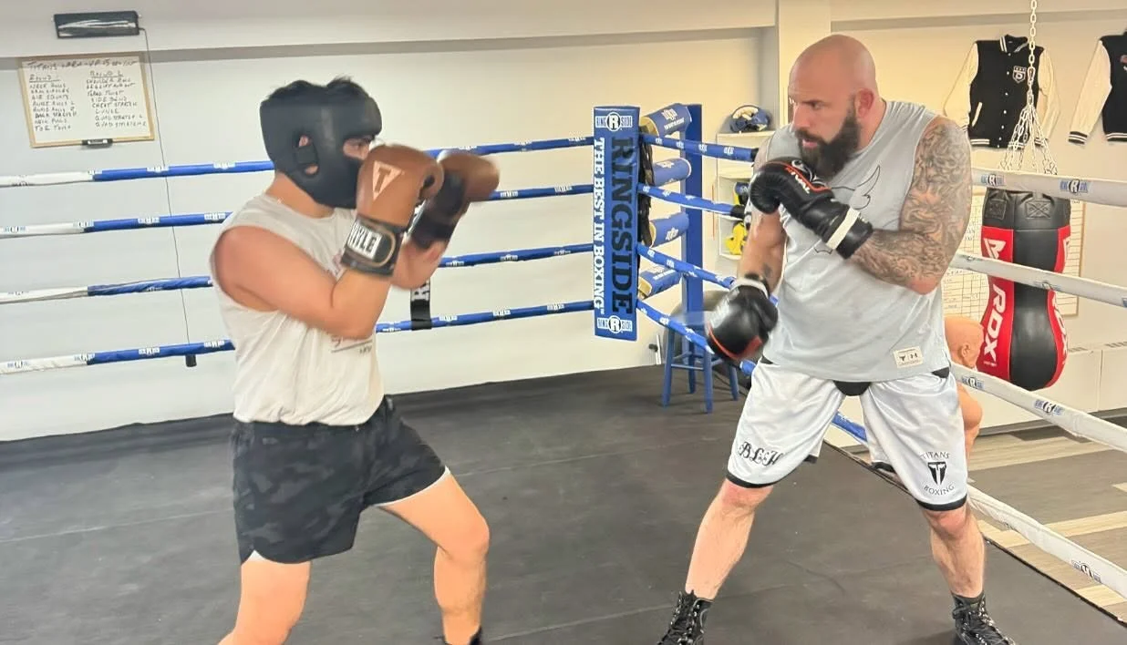 A young boxer wearing a black headgear and camo shorts practicing boxing moves with an adult trainer in a boxing ring. The trainer has a beard and is in a fighting stance, wearing boxing gloves and gray athletic clothing. Equipment and jackets are vi