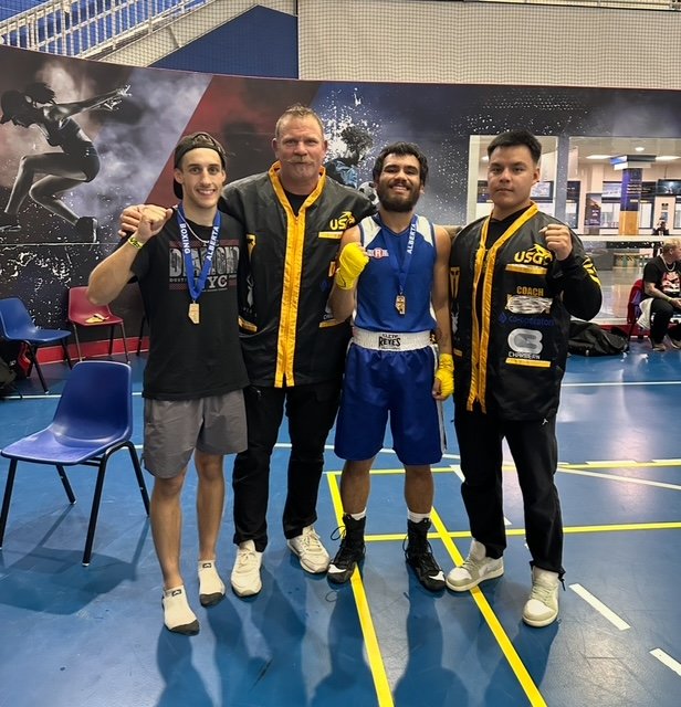 Four men in athletic attire, including a boxer in blue shorts, posing for a photo inside a sports venue. Three men are in black and yellow jackets, likely coaches or officials, while the boxer is in the center with yellow boxing gloves. One is wearin