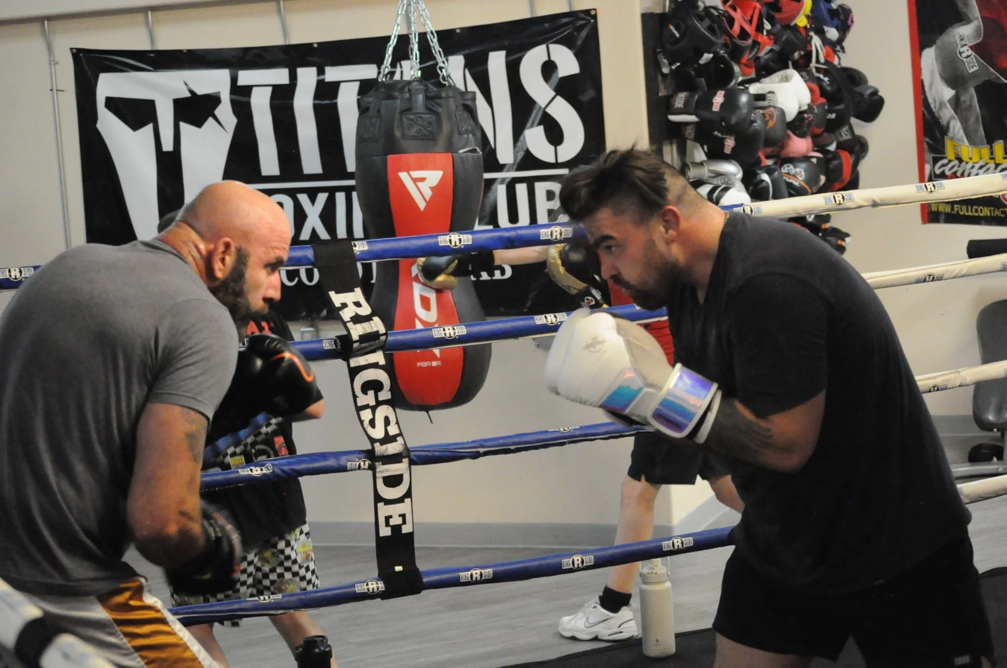 Two men training in a boxing gym, wearing boxing gloves and facing off with their gloves raised, with boxing ring ropes and equipment in the background.