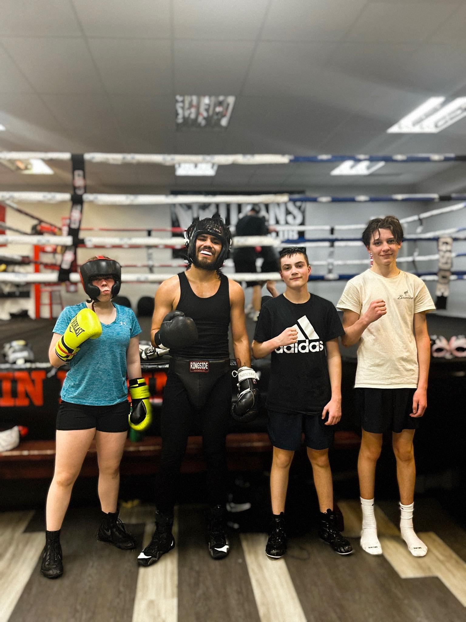 Four people in boxing gear and casual clothes posing in a boxing gym, with a boxing ring in the background.