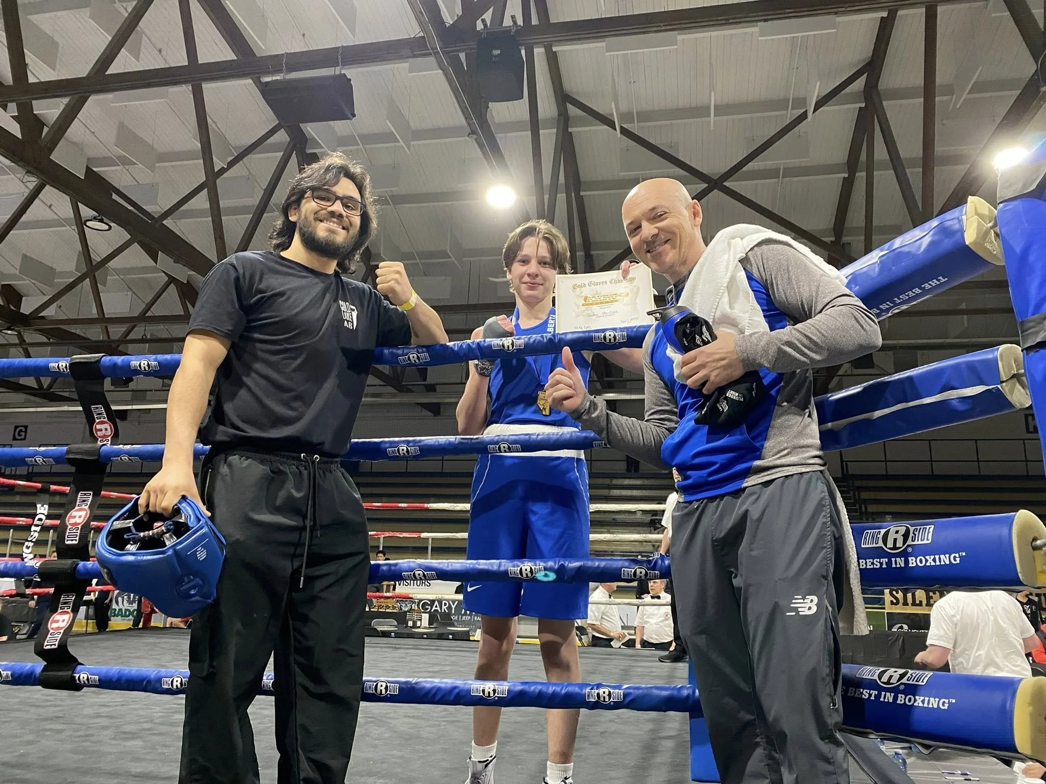 Three people in boxing ring celebrating; one woman holding a certificate, two men posing with fists, all smiling.