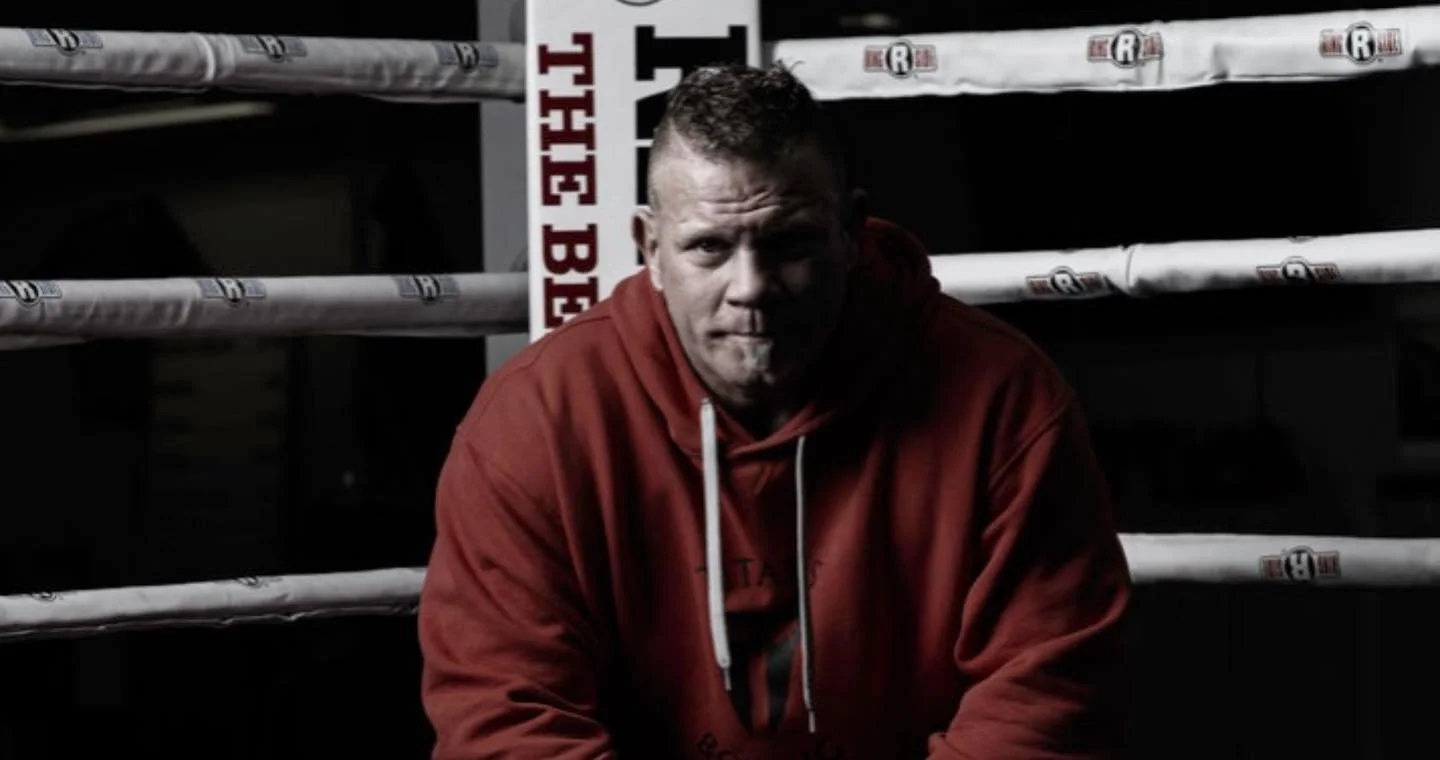 A man wearing a red hoodie sitting in a boxing ring with dark background and bright overhead lights.