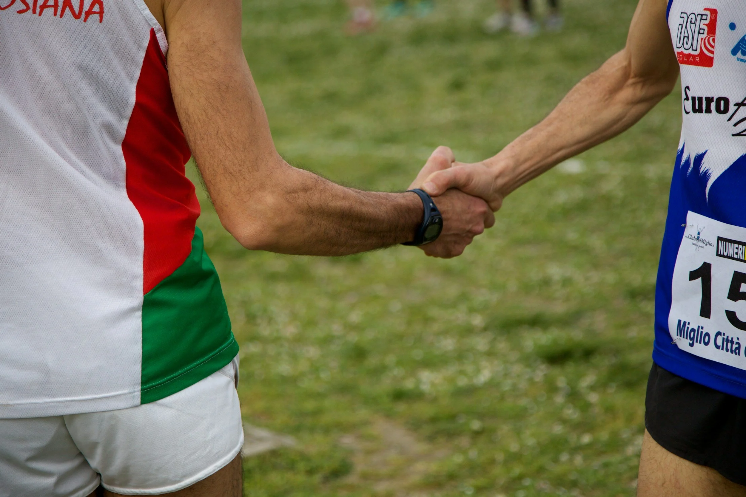 Two men shaking hands on a grassy field, one wearing a white sports uniform with red, green, and white colors, and the other wearing a white sports uniform with blue and red designs, and a numbered bib.