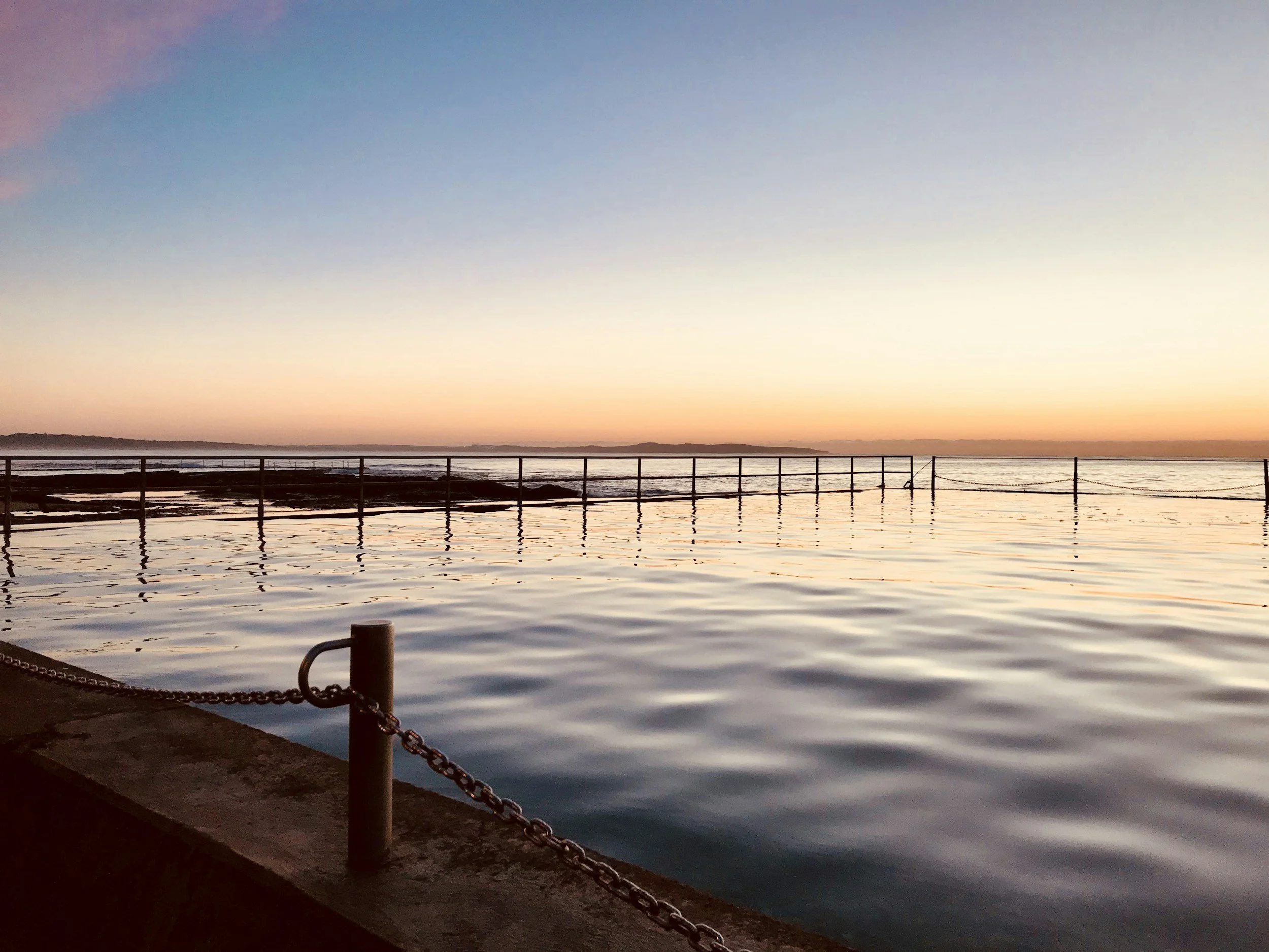 Sunset over the ocean with calm water, a pier or rail in the foreground, and distant land on the horizon.