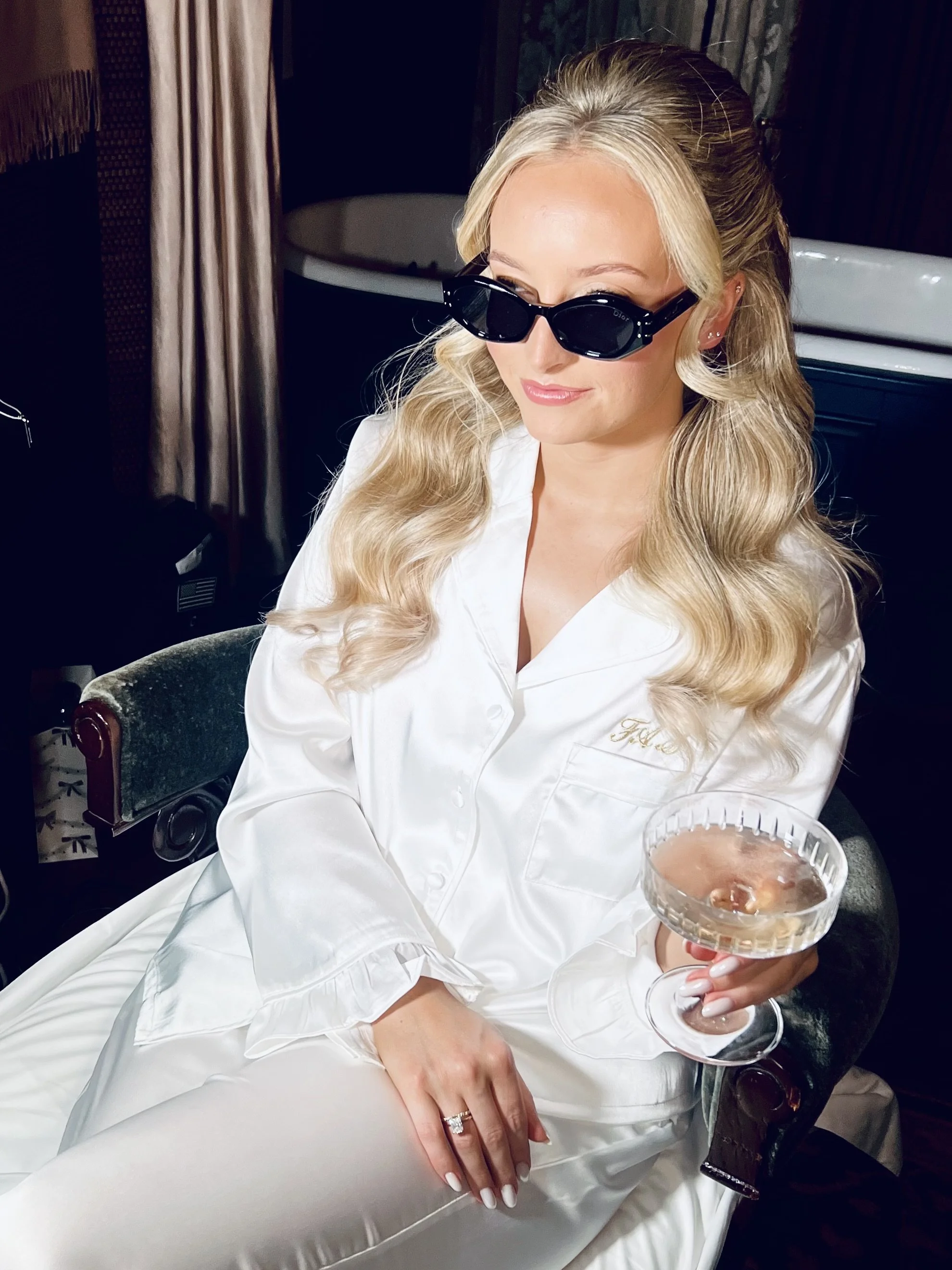 A glamorous bride to be with beautifully polished hair and makeup sits in her bridal pajamas sipping her champagne, ready to get dressed to Marry in a beautiful London hotel ceremony.