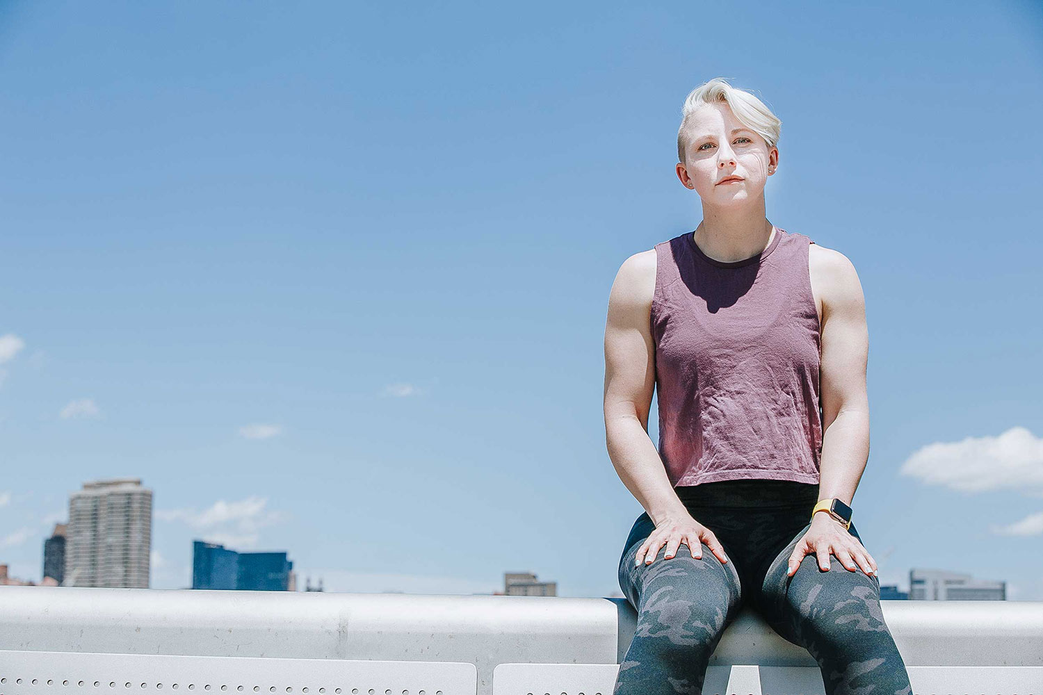 Megan in workout outfit, sitting beneath a blue sky, with hands on thighs.