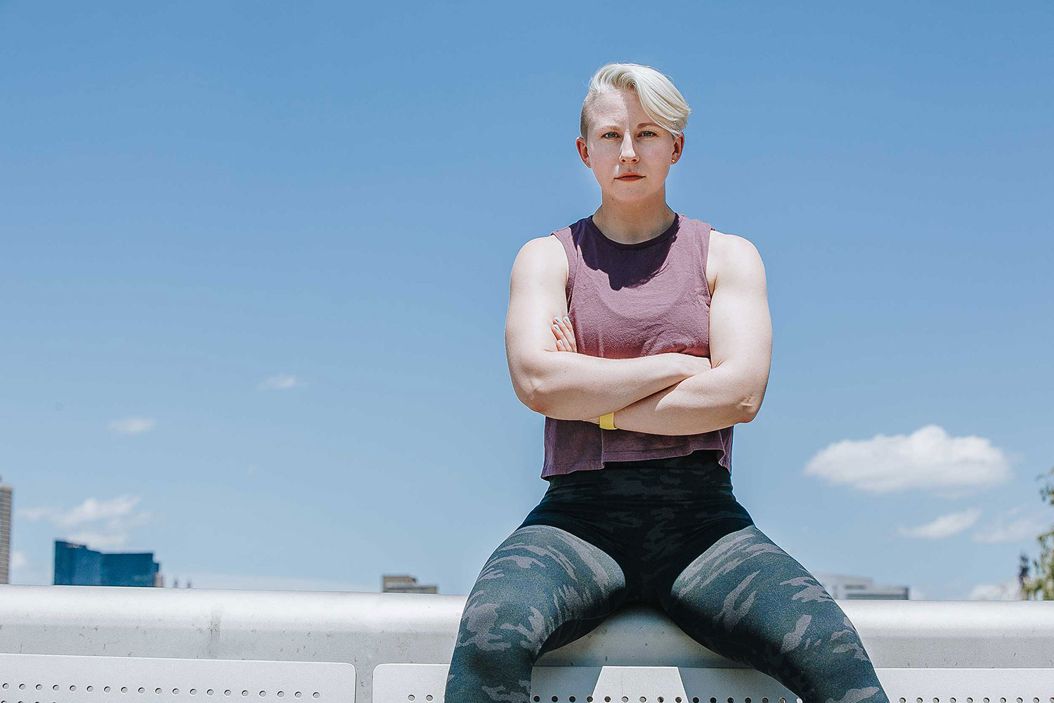 Megan in workout outfit, sitting beneath a blue sky, arms crossed at chest, looking tough.