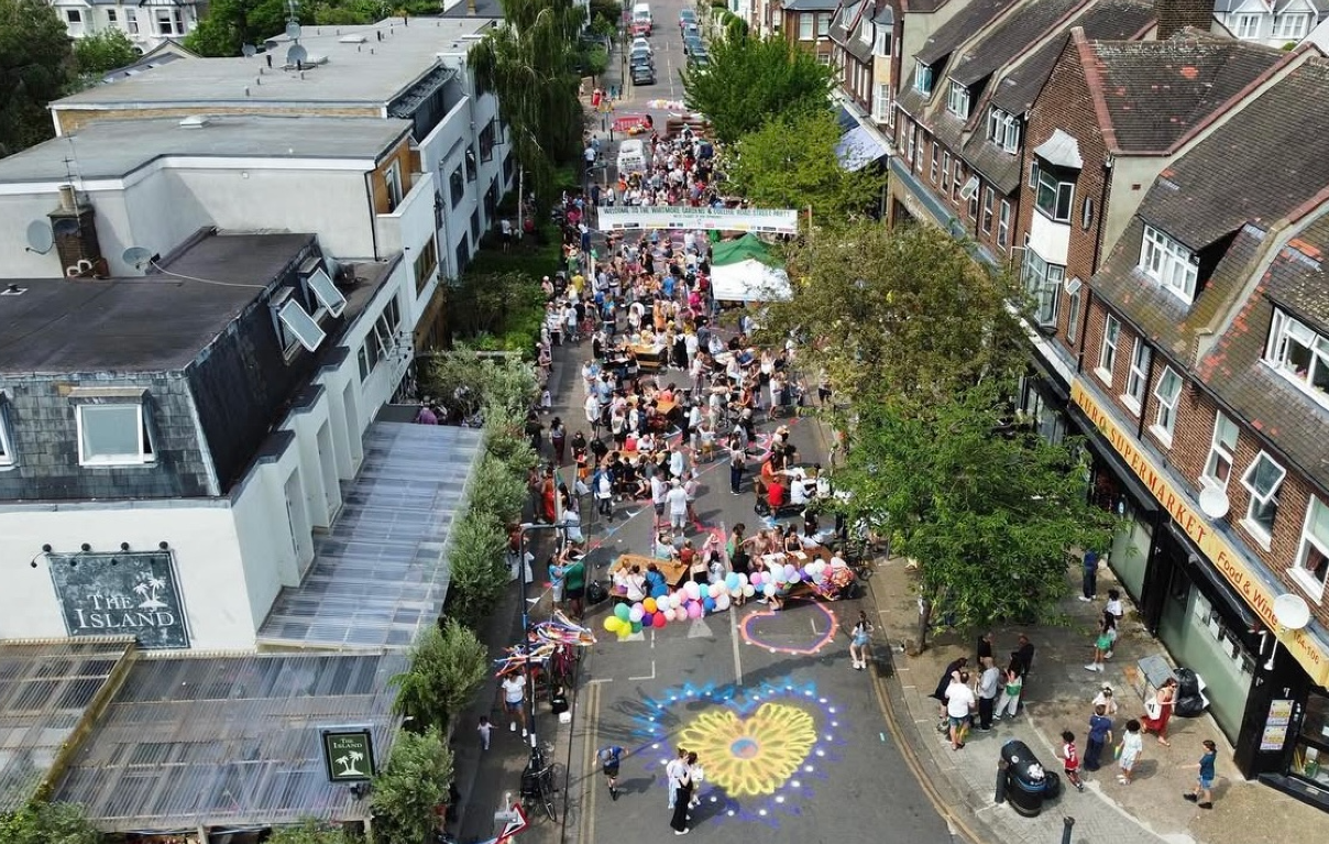 A street fair with a crowd of people walking along a street, vendors with booths, colorful balloons, and chalk art on the road, taken from an aerial view.