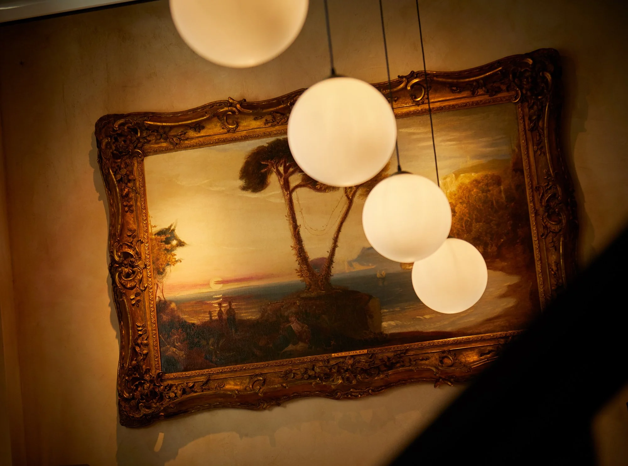 A framed landscape painting of a sunset scene with trees and water, illuminated by four round hanging light fixtures in a dimly lit room.
