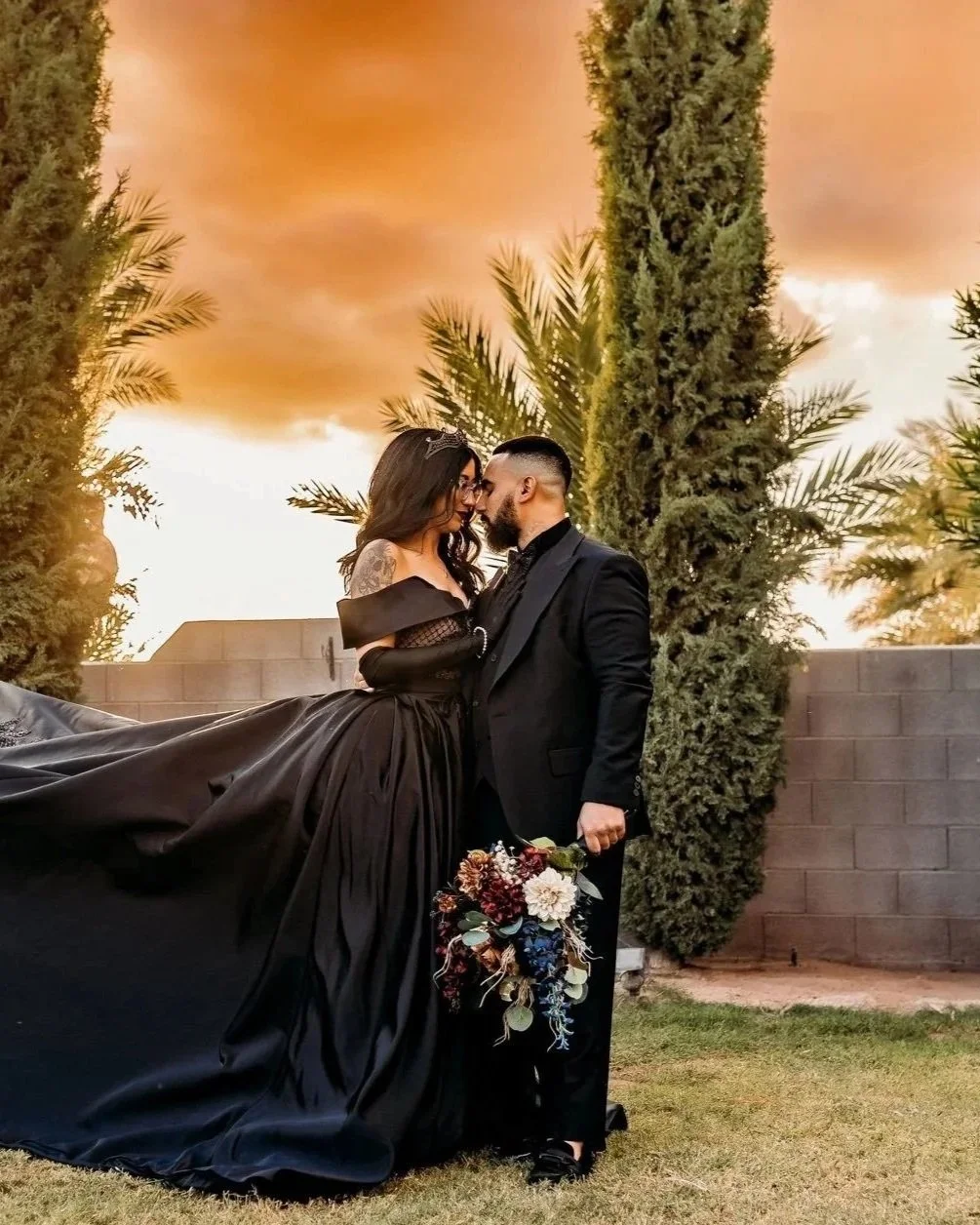 A couple dressed in formal wedding attire, standing close together outdoors at sunset, with a bride holding a bouquet of flowers and a groom in a black suit, surrounded by tall green trees and a brick wall.