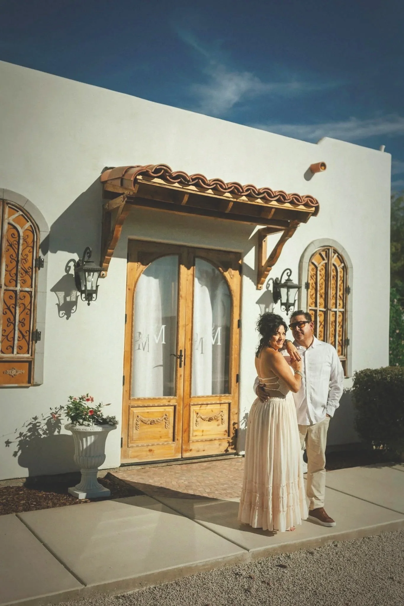 A man and woman standing together outside in front of a white building with wooden doors and windows. The woman is wearing a long, cream-colored dress and the man is wearing a white shirt and pants. The sunlight casts shadows on the wall behind them, and there is a white decorative urn with flowers near the building.