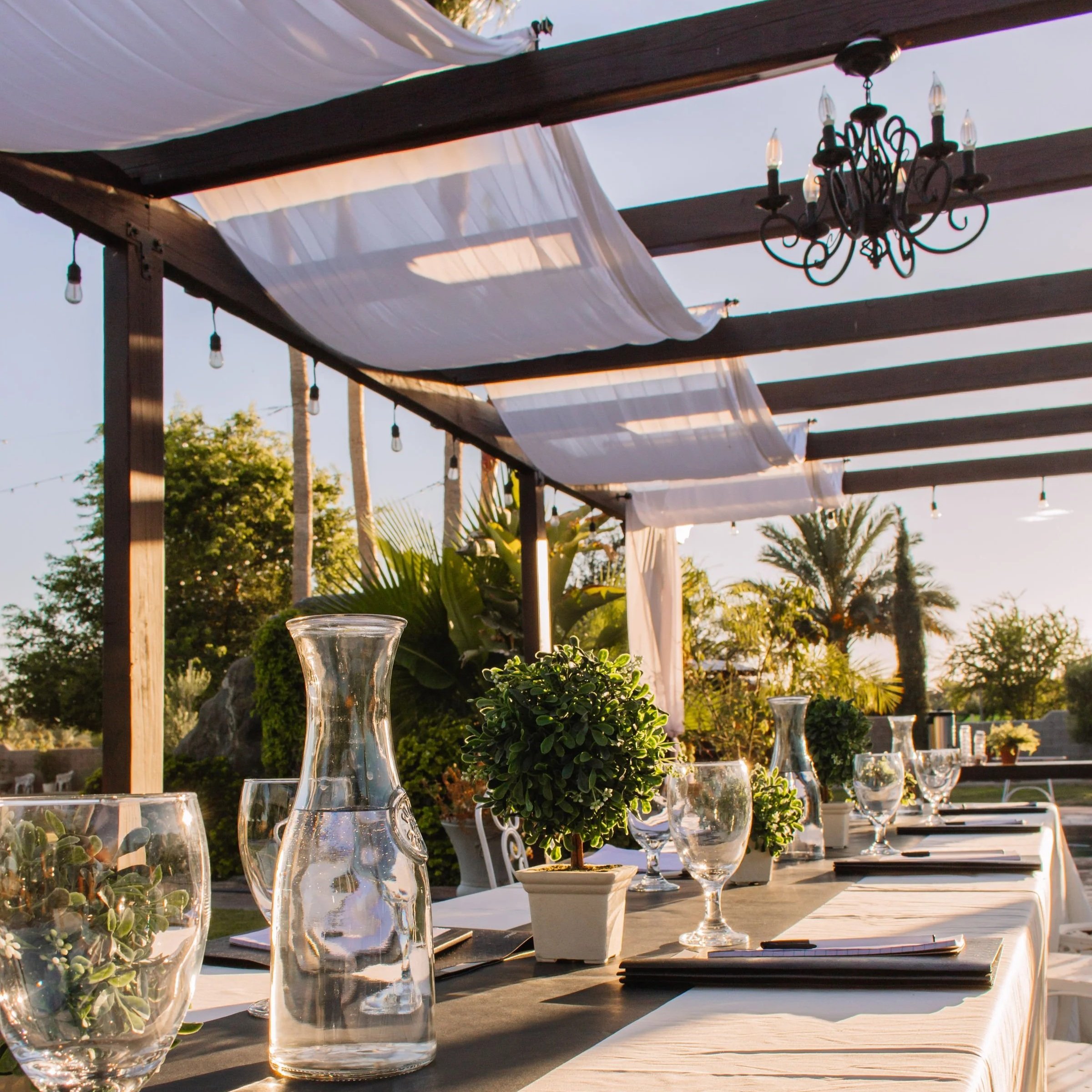 Outdoor table set with glass carafes, potted plants, and tableware under a wooden pergola with a chandelier, during sunset.