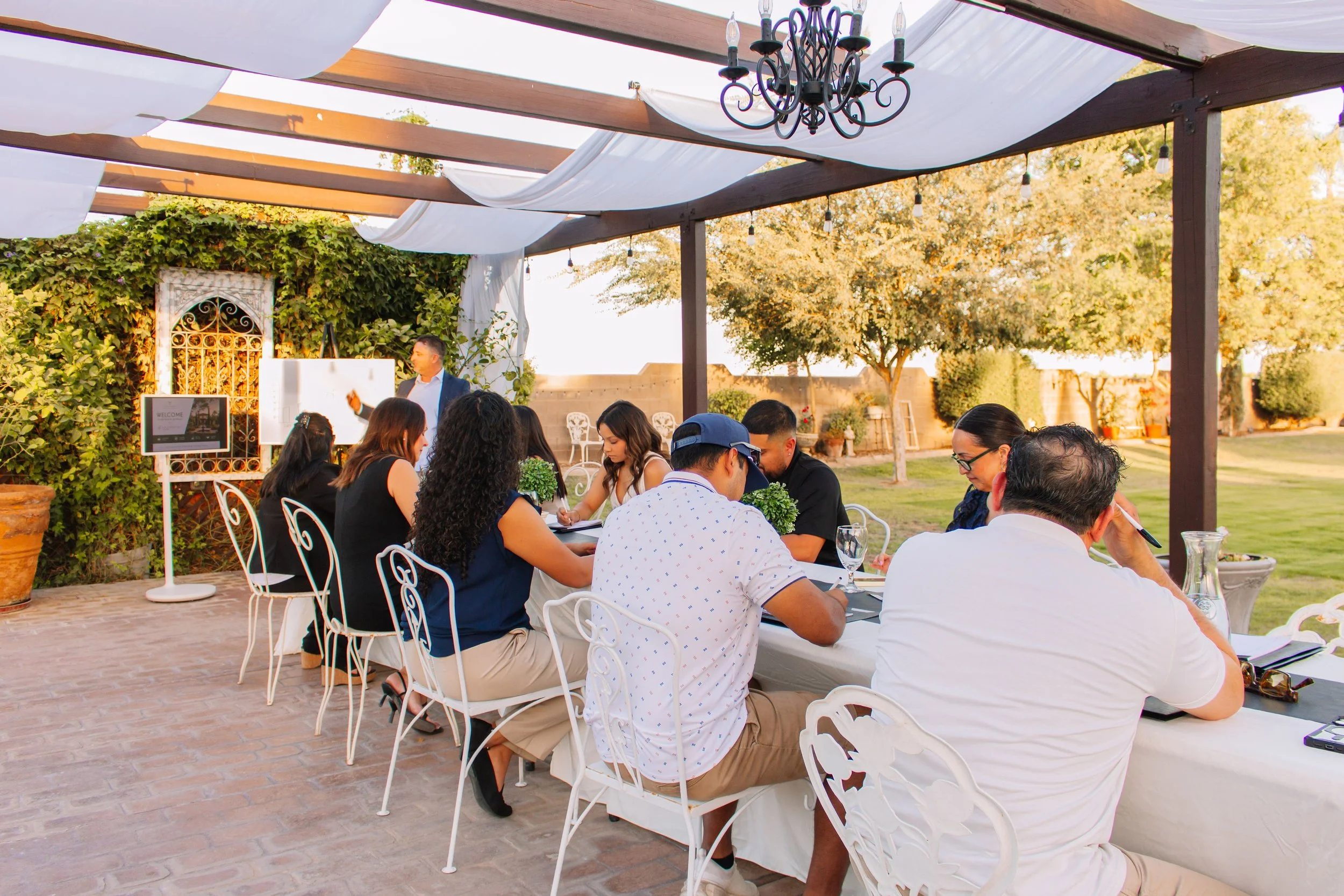 People sitting at a long outdoor table under a pergola with white fabric, with a man standing and giving a presentation or speech in the background, during daytime in a garden setting.