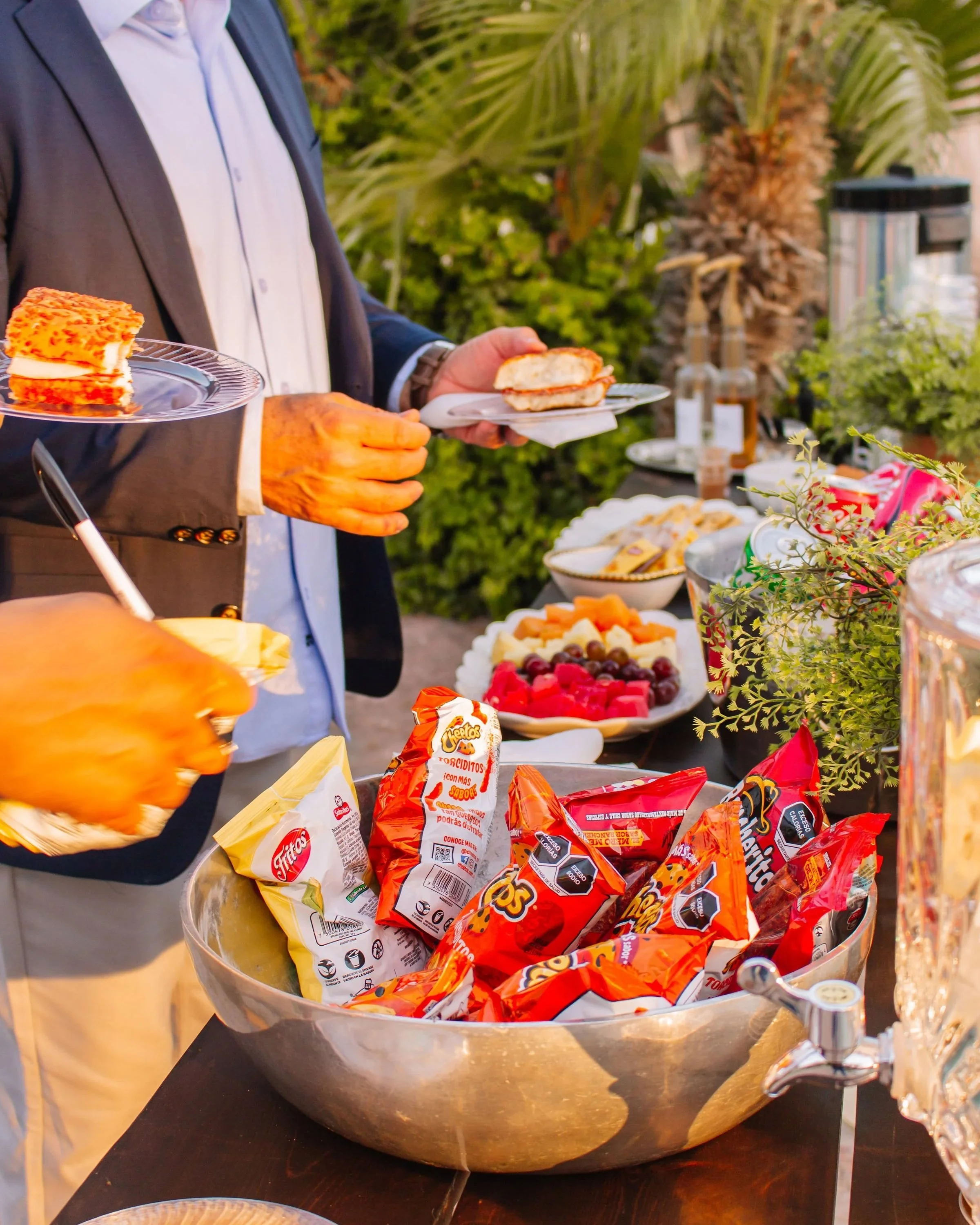 Person in a suit serving snacks at an outdoor party with a large bowl of chips and bowls of fruit, desserts, and condiments on a table in background, surrounded by greenery.