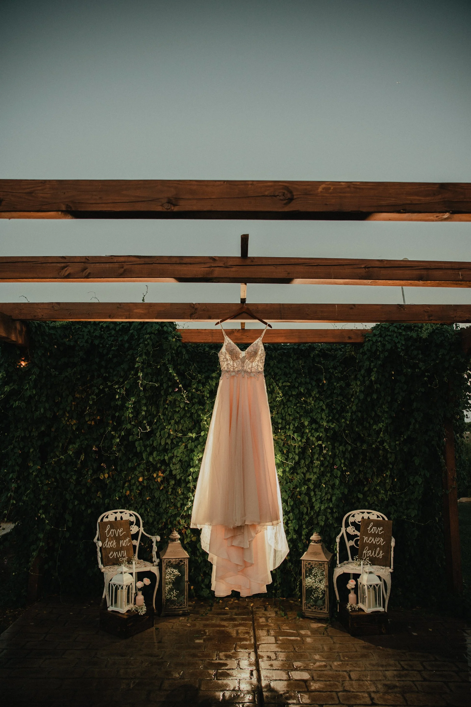 A peach-colored wedding dress hanging from a wooden beam against a green leafy background, with two white chairs and decorative signs that say "love does not envy" and "love never fails" on either side.