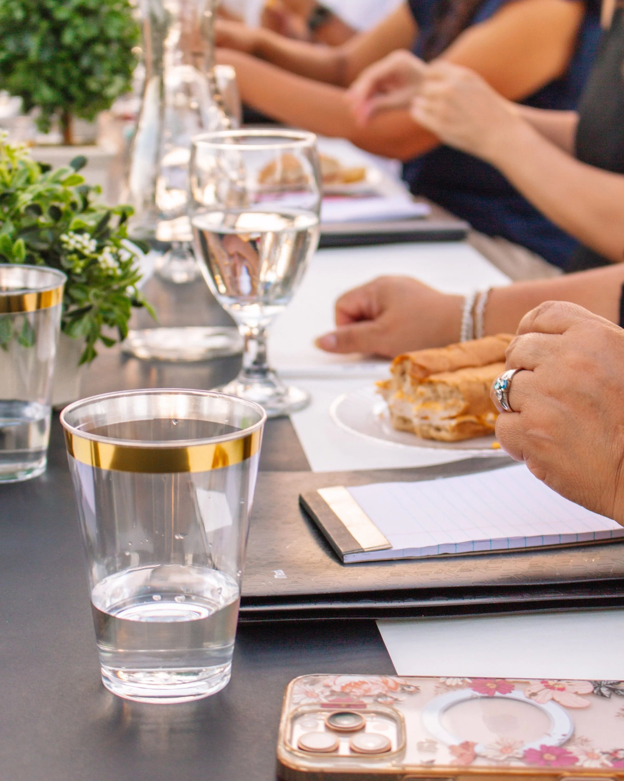 Close-up of a glass of water and various objects on a black table, including a potted plant, a notebook, and a person's hand holding a snack, with people sitting in the background.