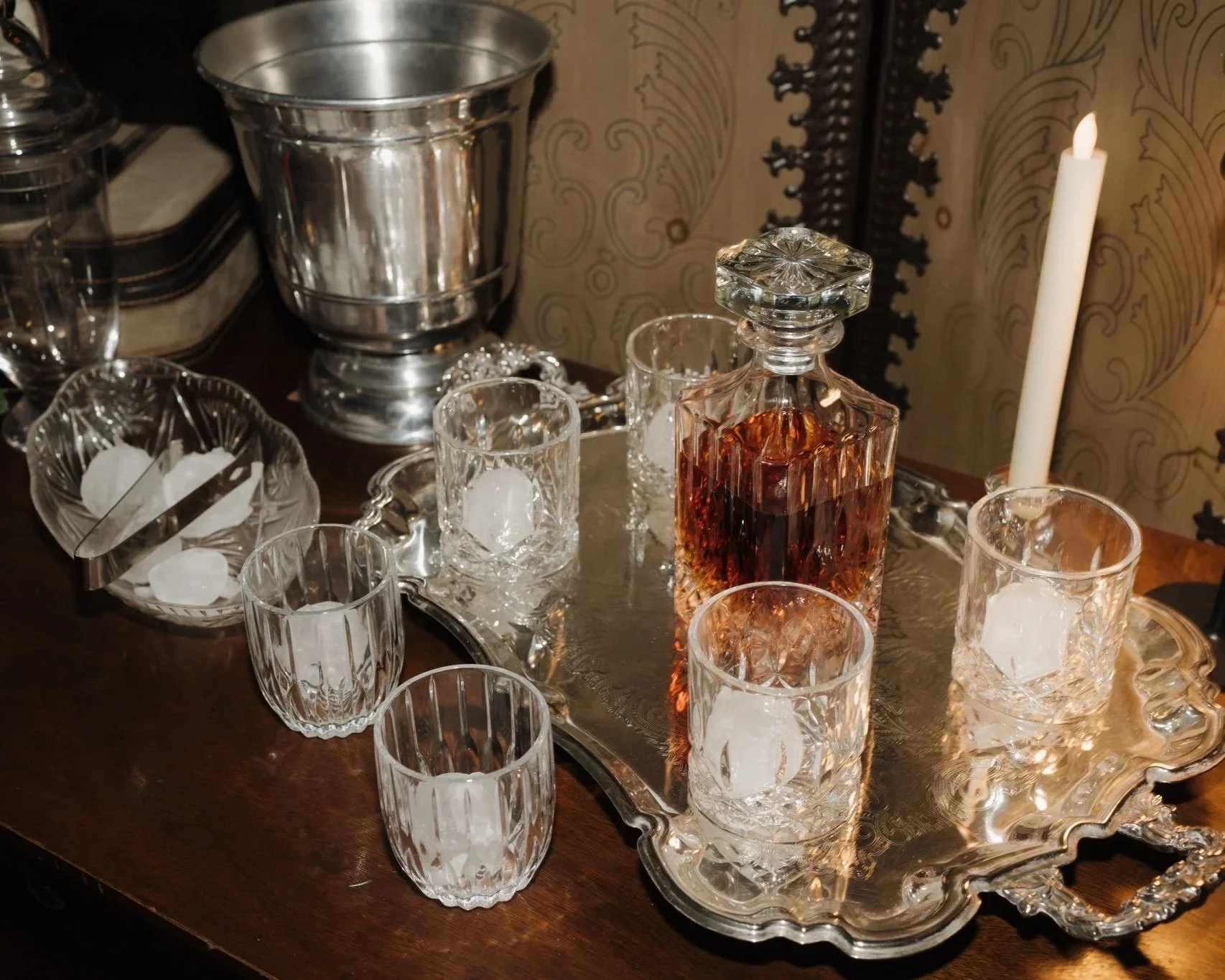 A collection of glassware including a decanter with amber-colored liquid, several glasses with ice, a candle, and silver serving items on a silver tray and tray with an ornate edge, set on a dark wooden table against a decorative wall.