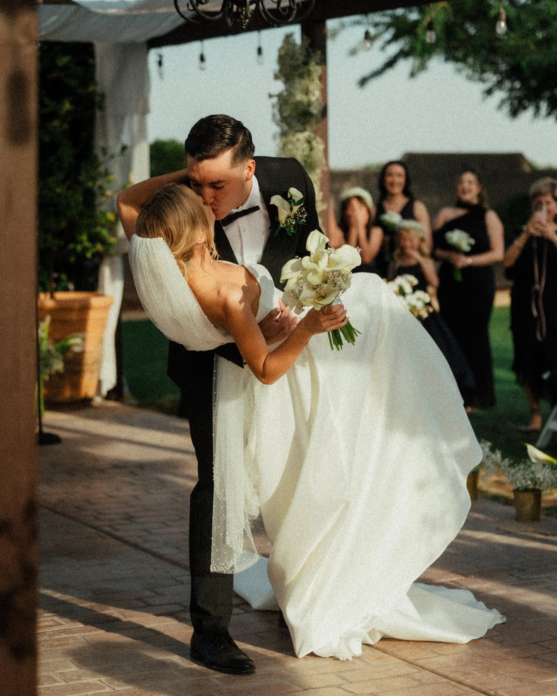 A groom in a tuxedo and a bride in a wedding dress sharing a kiss at their wedding reception outdoors during sunset.