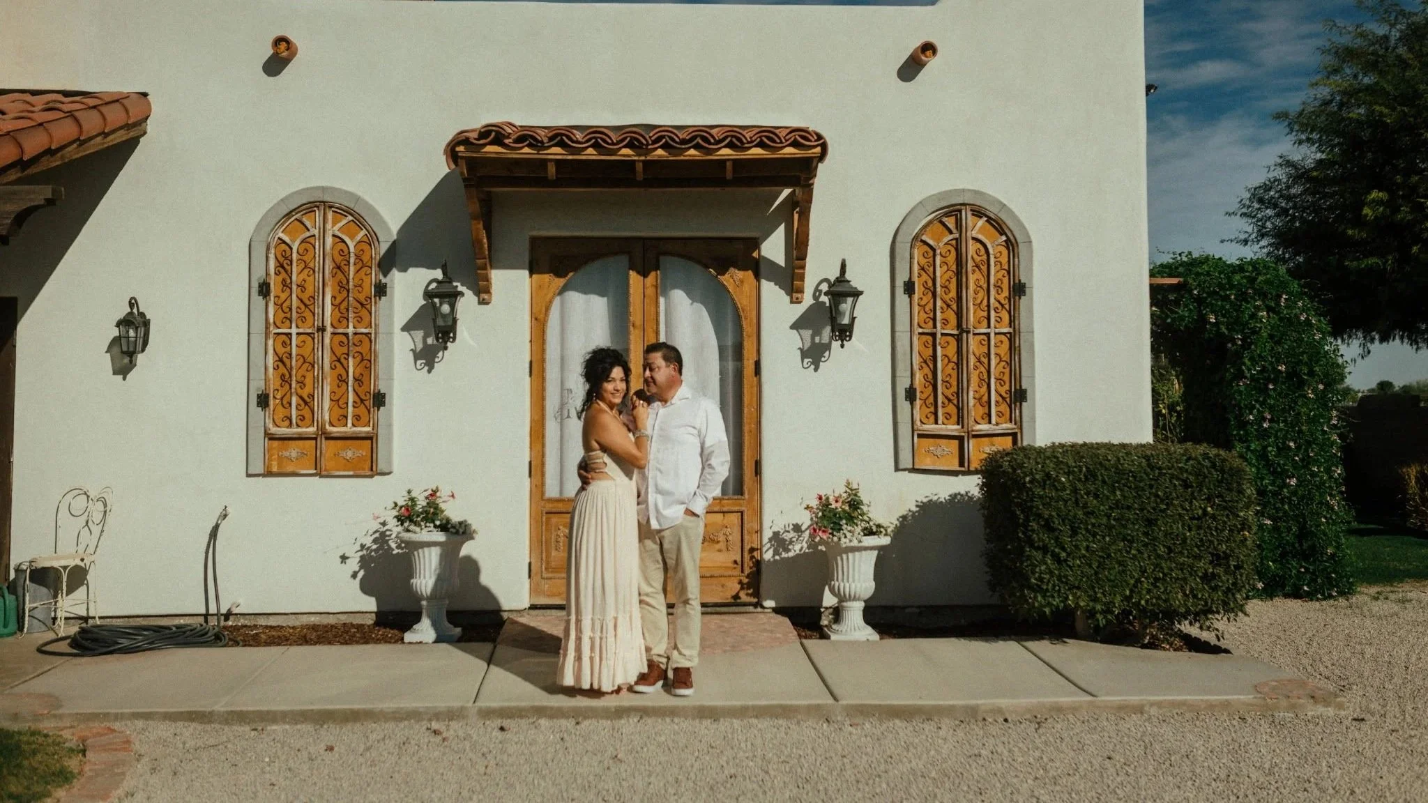 A couple stands close together in front of a white house with two arched windows with decorative wrought iron grilles and a wooden door. The woman is on the left, wearing a long, flowing dress, and the man is on the right, wearing a white shirt and beige pants. They are smiling and looking at each other. The house has a small porch with a wooden awning. There are two wall-mounted lanterns on each side of the door and potted plants with flowers on either side of the entrance. A tree and bushes are visible to the right of the house, and the sky is partly cloudy.