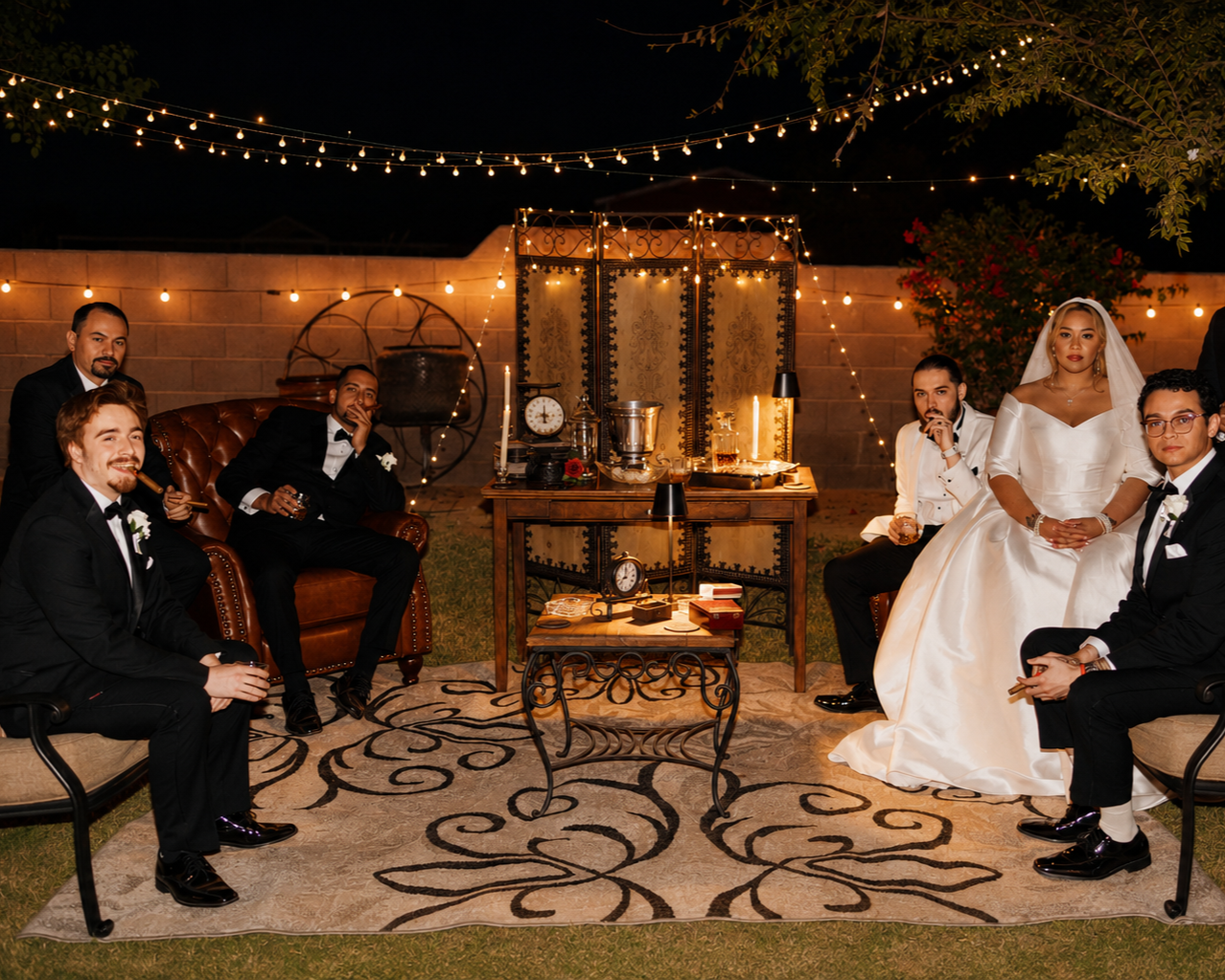 Group of people at a wedding reception outdoors at night with string lights, vintage furniture, and decorative items on display.