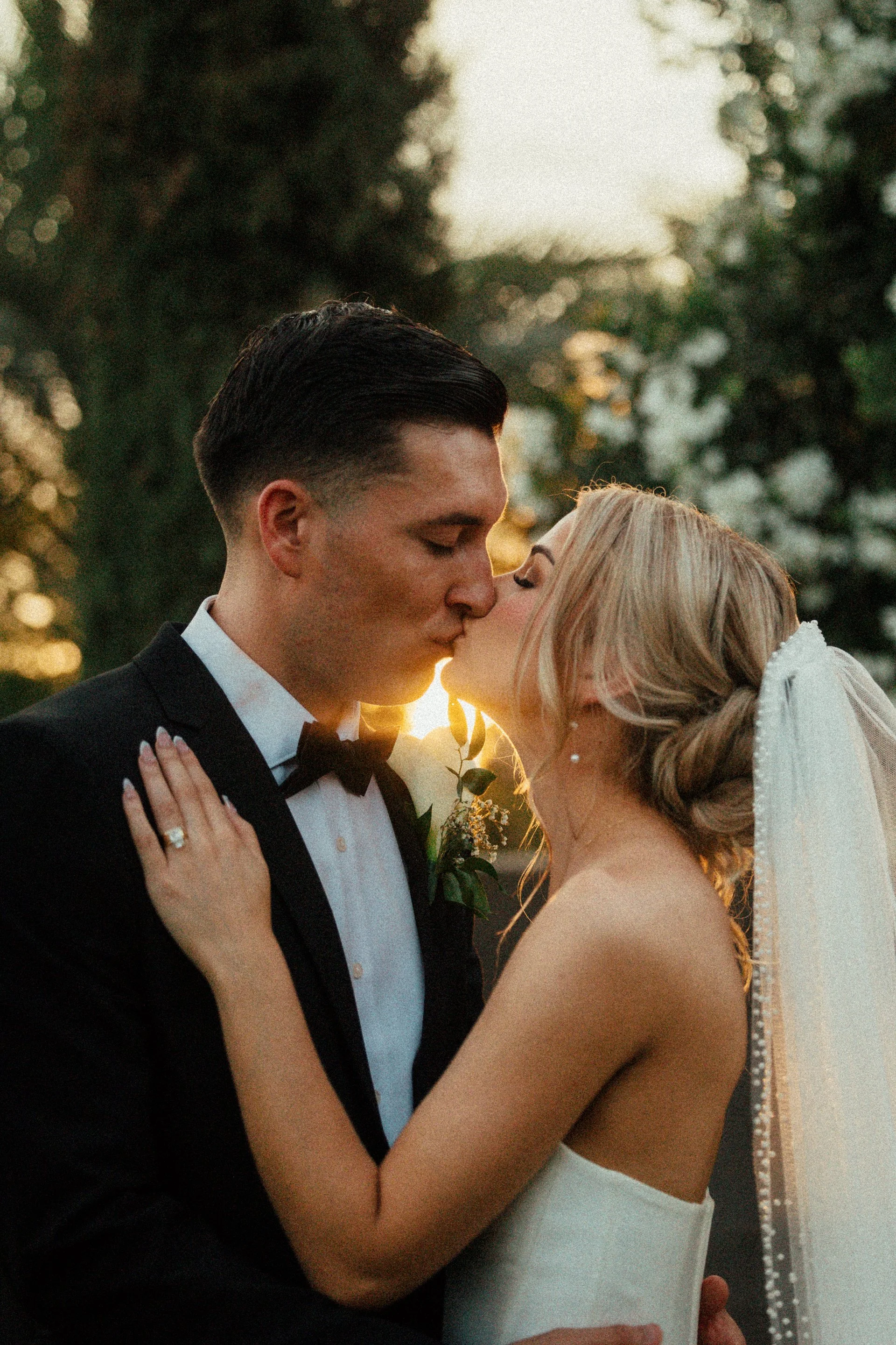 A bride and groom sharing a kiss outdoors during sunset, with trees in the background.