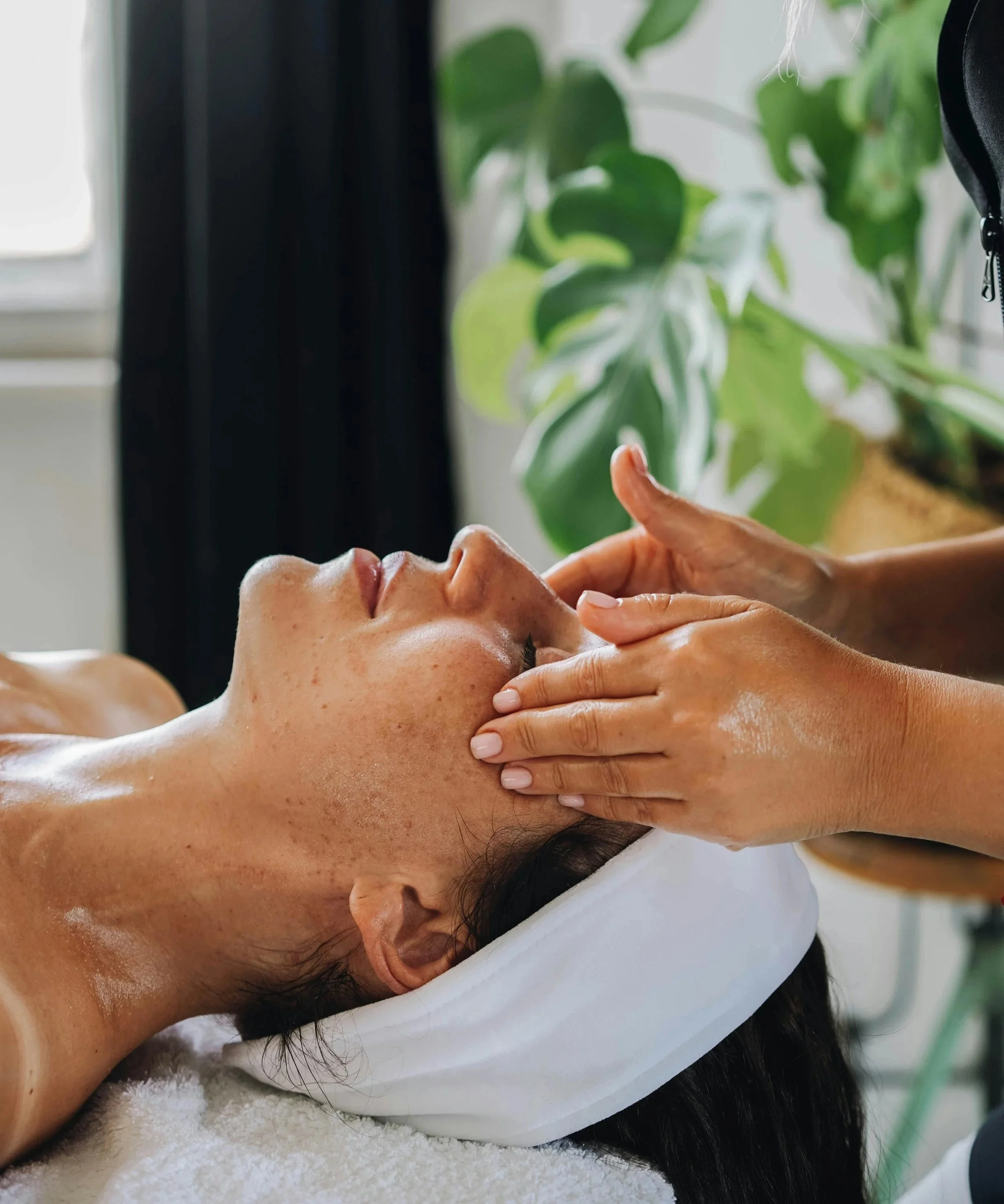 A woman receiving a facial massage at a spa, lying on a treatment bed with a white towel wrapped around her hair and a white headband, as a skincare professional gently massages her face.
