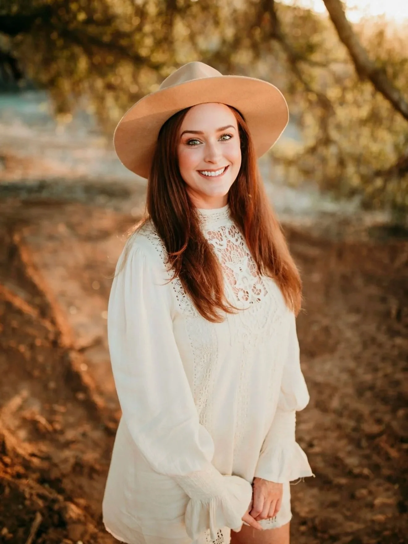 A woman with long red hair, smiling and wearing a wide-brimmed beige hat and a white lace dress, standing outdoors in a natural setting during sunset.