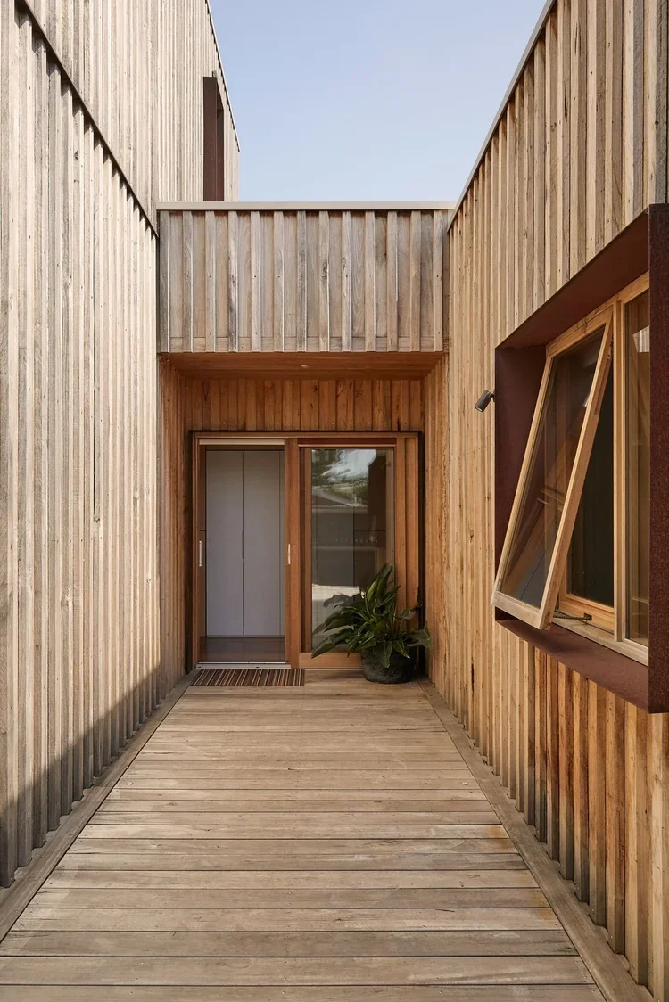 Exterior view of a modern wooden building with a wooden deck, a glass sliding door, a potted plant, and an open window.