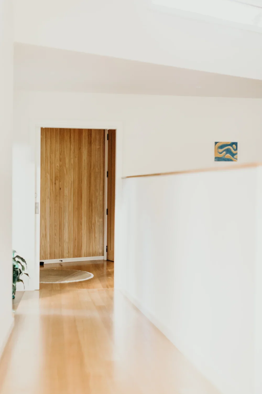 Interior hallway with wooden flooring, white walls, and a wooden door at the end. A small abstract artwork hangs on the right wall, and a round rug is visible on the floor near the door.