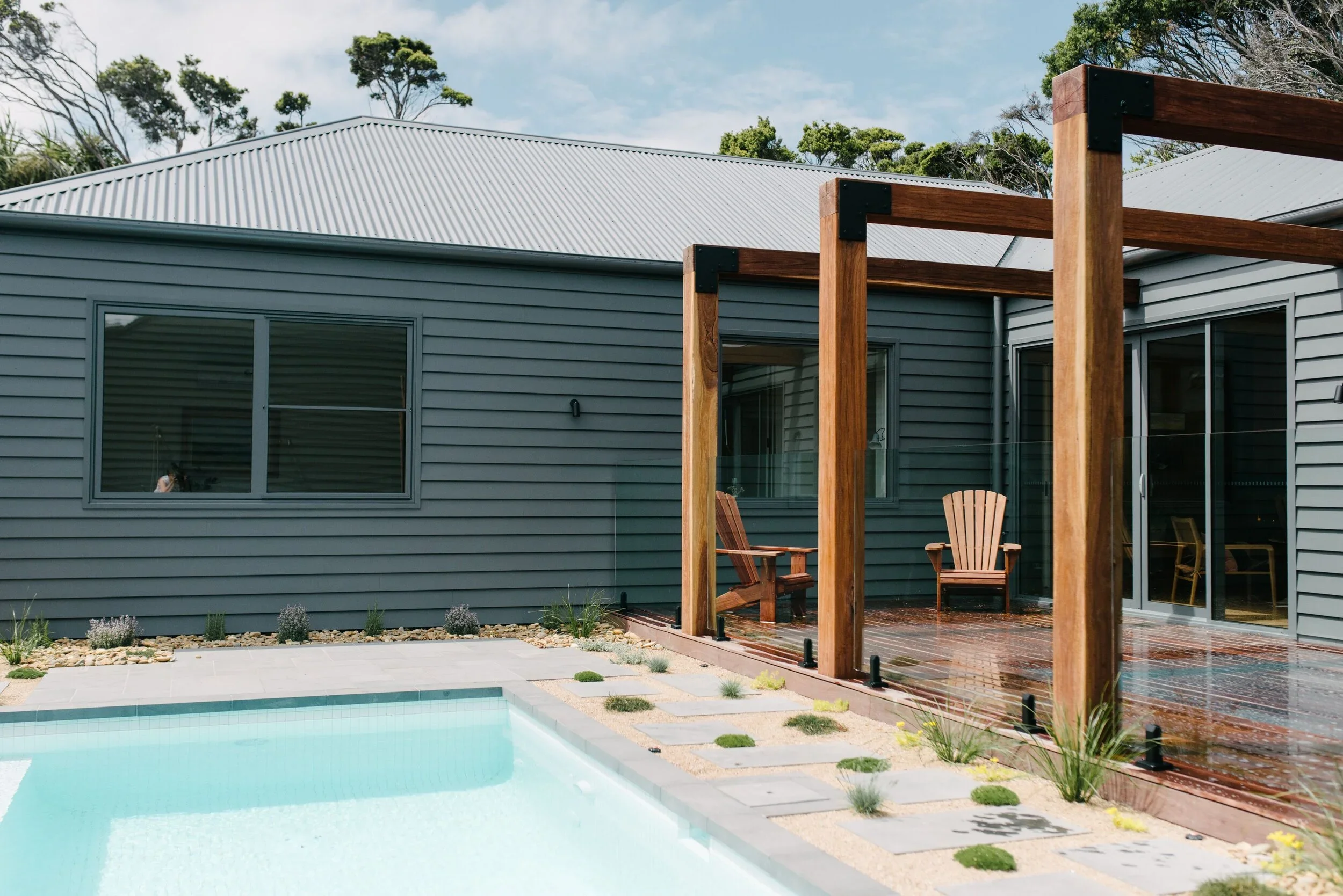 Modern house backyard featuring a swimming pool, wooden deck, and a shade structure with wooden beams, surrounded by trees and landscaping.