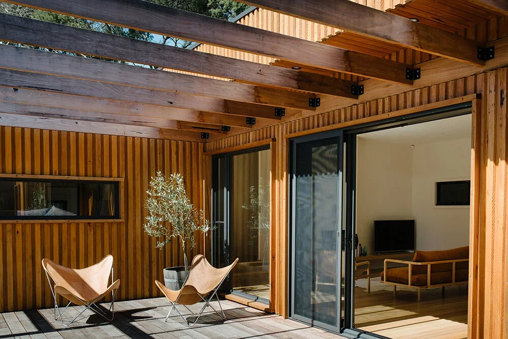 Outdoor patio area with wooden decking, two tan butterfly chairs, and a potted plant, adjacent to an interior room with a TV and tan sofa visible through sliding glass doors.