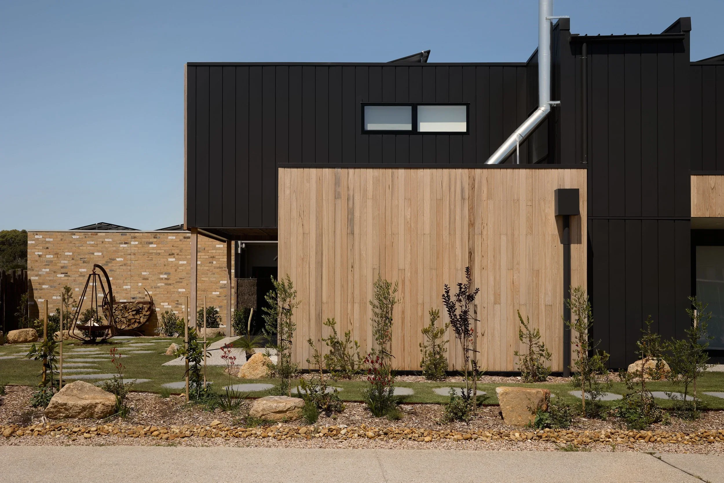 Modern house with black and wood exterior, small window, pipe on the wall, garden with flowers and rocks, gravel path, and a swing set in the yard.