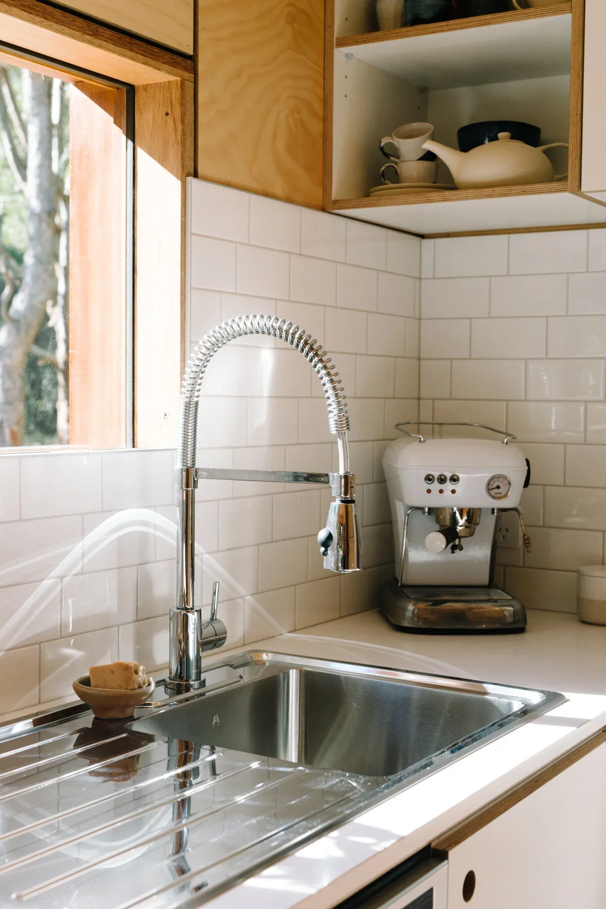 A kitchen sink with a chrome faucet, a soap dish with soap, and a coffee machine on a white countertop, with wooden cabinets and a window in the background.