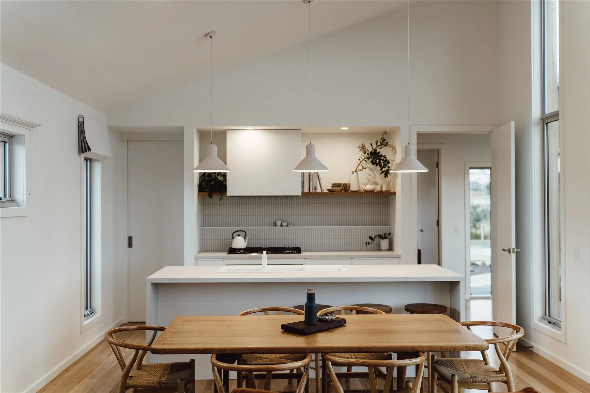 Modern kitchen with white cabinetry, pendant lights, a brick backsplash, and large windows, opening to a dining area with a wooden table and chairs.