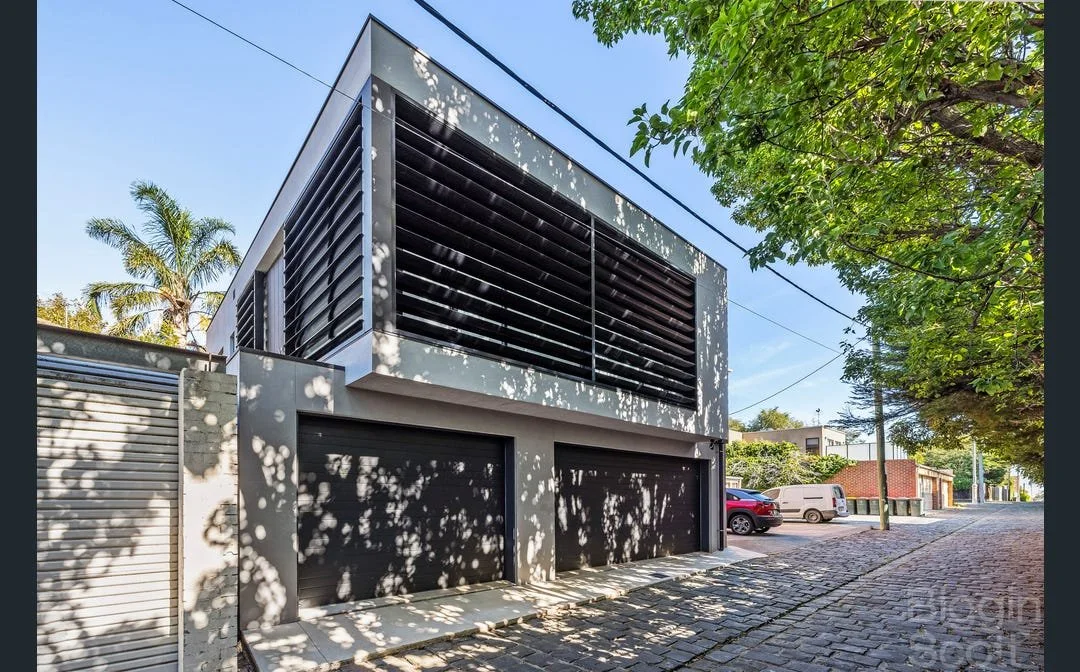 Modern two-story house with black window shutters and two black garage doors, on a tree-lined street with parked vehicles.