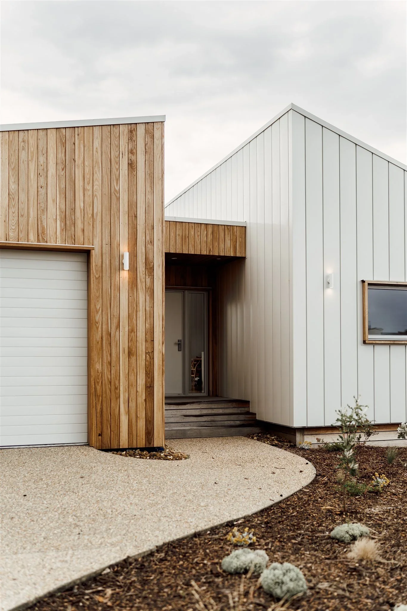 Modern house with a combination of white and wooden exterior, featuring a garage door and adjacent entryway, with landscaped front yard including small plants and mulch.