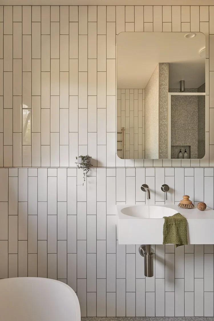 A modern bathroom with white rectangular tiles on the wall, a large mirror, a white wall-mounted sink with a green towel, a small decorative object, and a plant hanging on the wall.