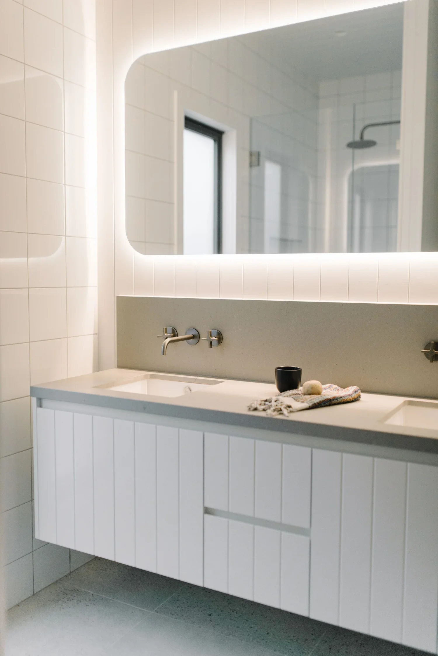 Modern bathroom sink with a large mirror backlit with LED lighting. The sink cabinet is white with shiplap paneling. On the countertop, there are two small objects, possibly soap or skincare items, along with a black cup or container, and a small tow