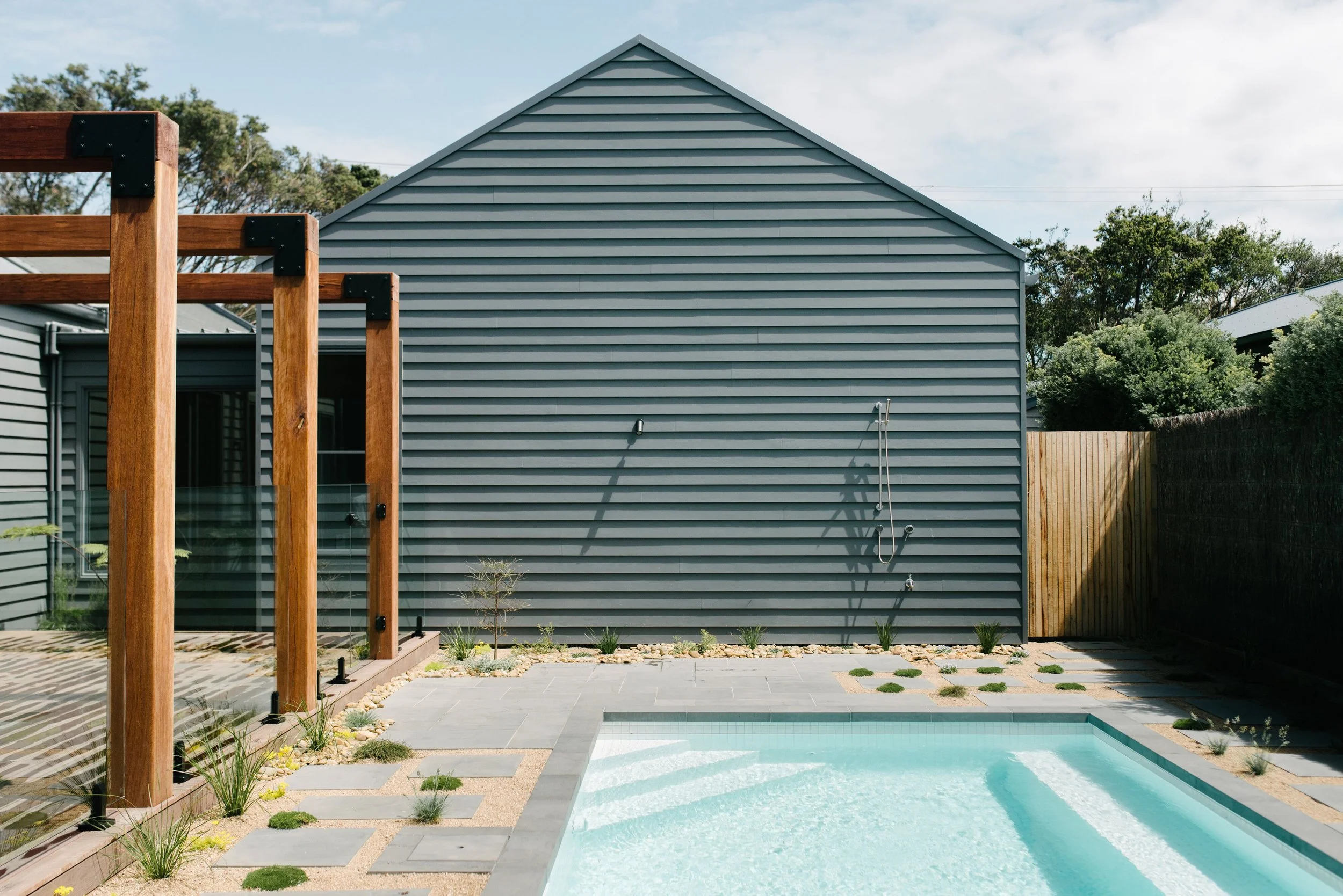 A backyard with a small swimming pool, a gray house with siding, a wooden fence, and a wooden pergola frame, with trees in the background.