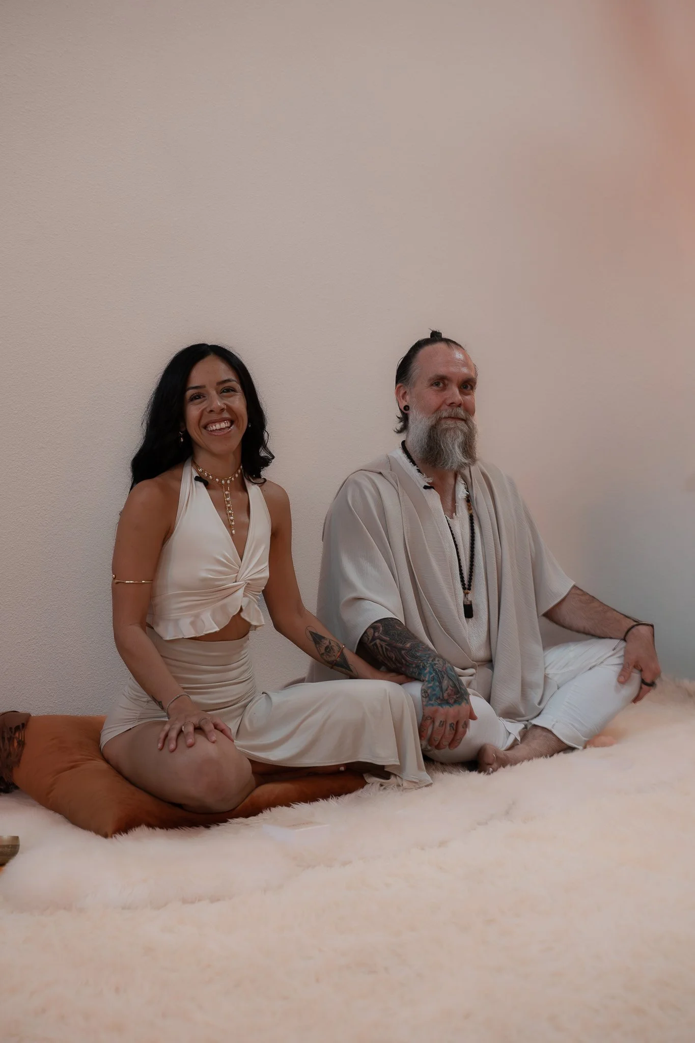A woman and a man sitting cross-legged on a plush fur rug against a plain wall. The woman is smiling, wearing a white dress, with creative jewelry, while the man has a long beard, tattoos, earrings, and is dressed in white spiritual attire.
