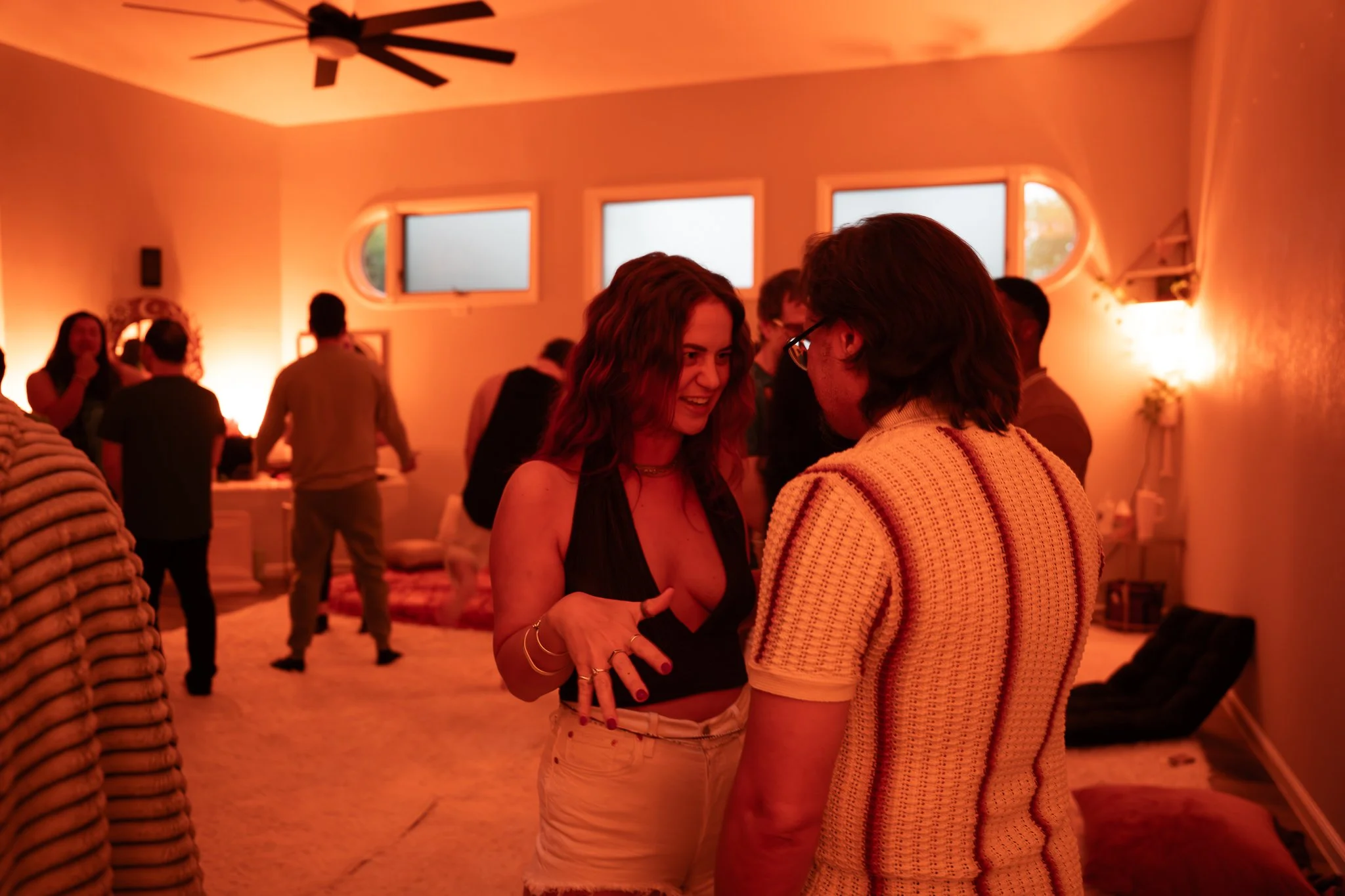 People dancing and socializing in a warmly lit room with orange and pink ambient lighting, rectangular and oval windows, and a ceiling fan.