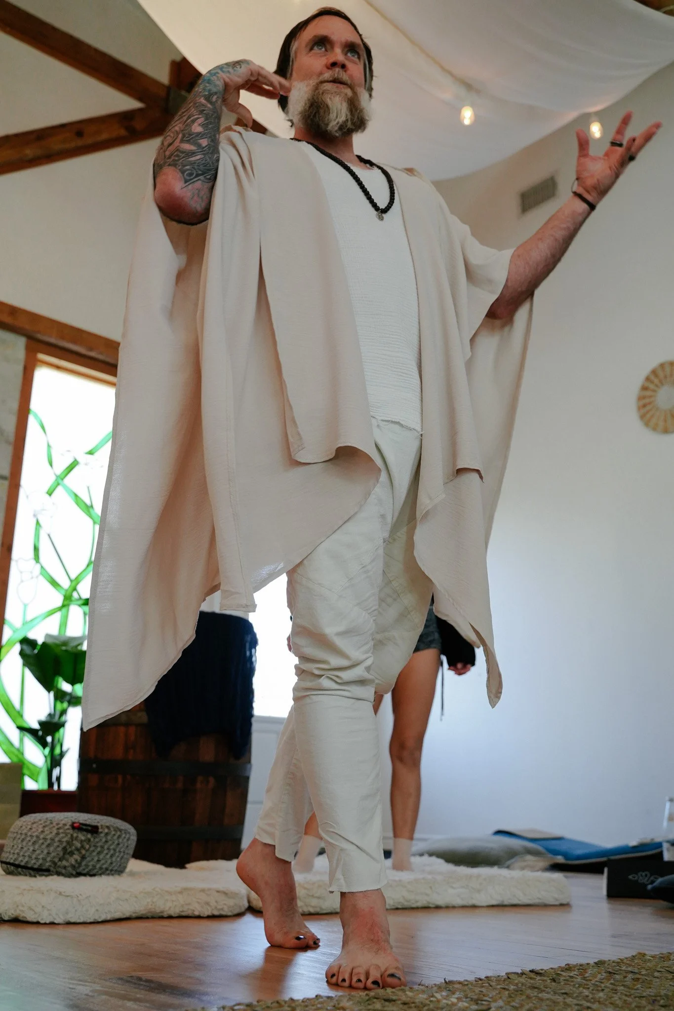 A man with tattoos on his left arm, a beard, and dressed in white, is inside a room with wooden beams, a tapestry on the ceiling, and a woven basket on a cushion on the floor. He appears to be in a yoga or meditation pose, standing barefoot on a wooden floor.