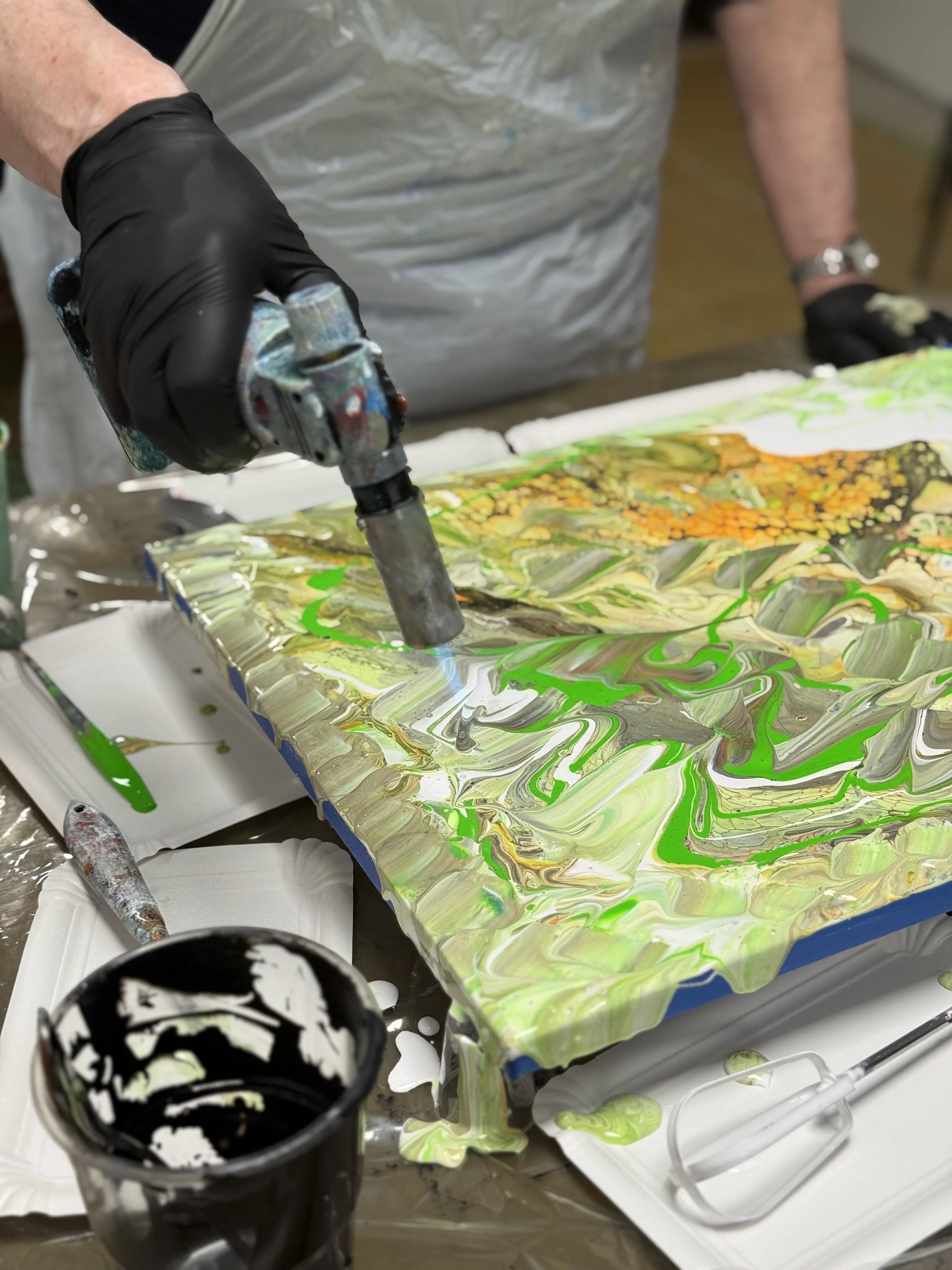 En person bruger en varmepistol til at skabe resin kunst på en arbejdsoverflade, omgivet af farvede resin materialer.