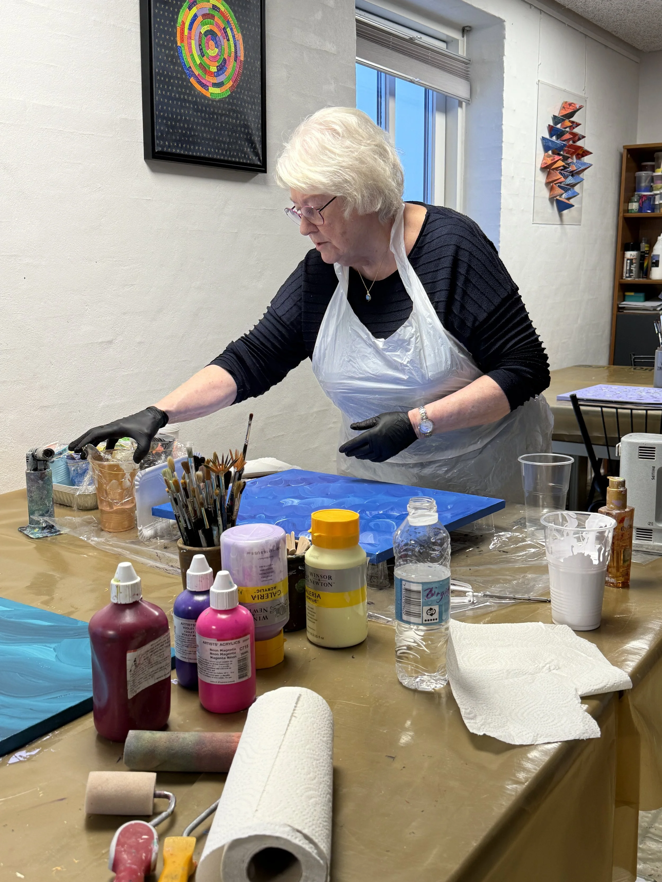 En ældre kvinde malearer på et bord med forskellige kunstmaterialer i et kunststudie.